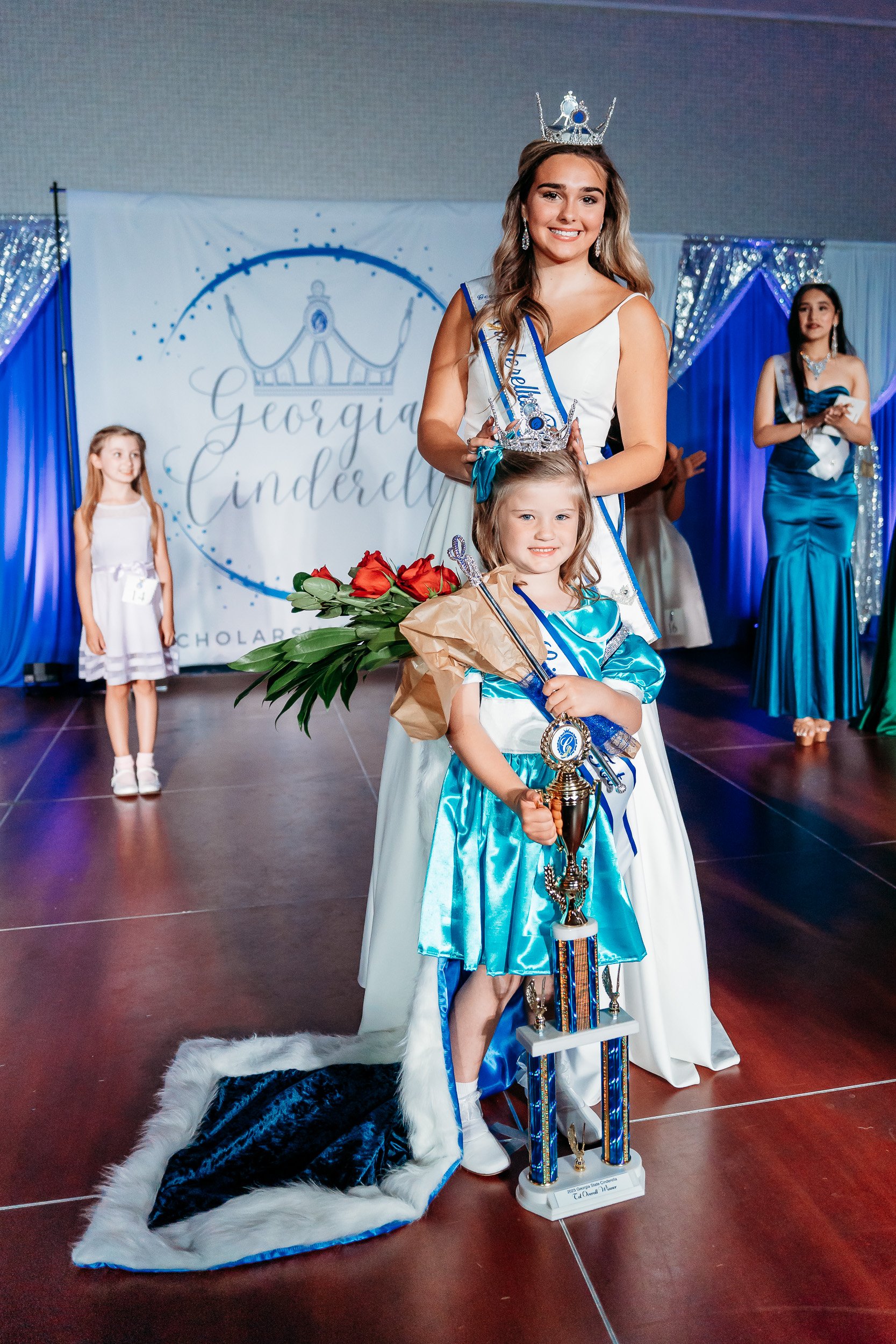 Beauty pageant scene with young girls dressed in gowns and crowns, one girl in blue holding a large trophy and bouquet of roses, another girl in white crowned and smiling, standing on stage with a sign reading 'Georgia Cinderella' in the background.