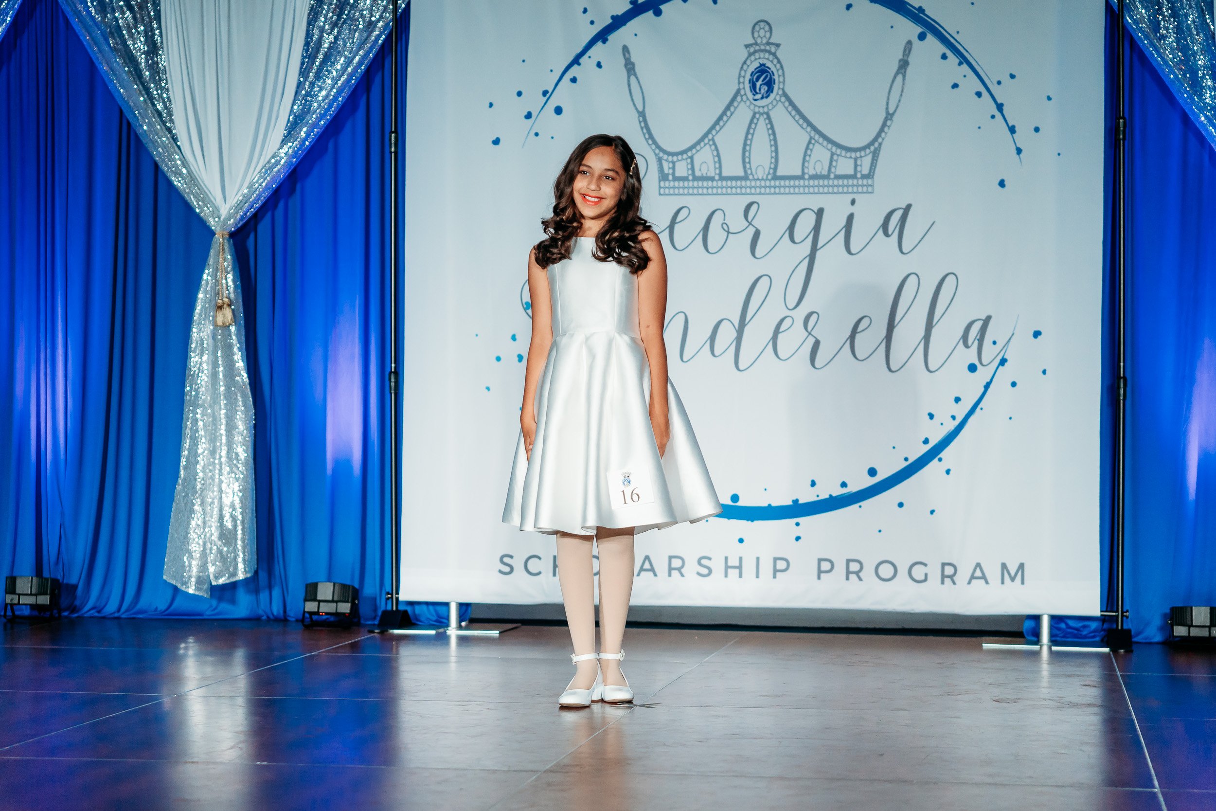 Young girl standing on stage at scholarship program event, wearing a silver dress and white shoes, with a bright smile. The backdrop reads 'Georgia Cinderella Scholarship Program' with a crown illustration.