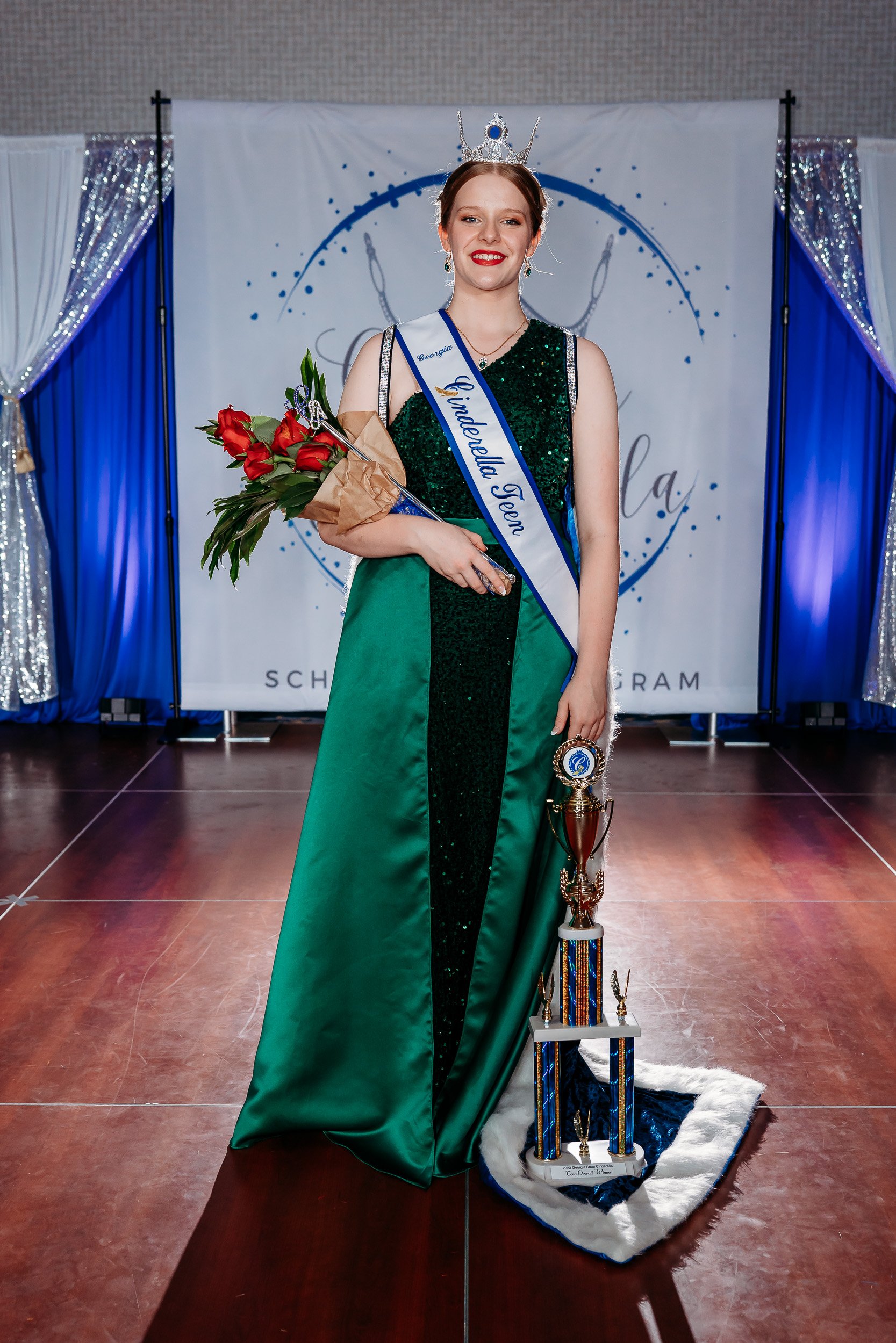 A young woman wearing a crown and sash reading 'Georgia Underdelia Teen,' in a green and black dress, holding a bouquet of red roses and a trophy, standing in front of a decorated backdrop.