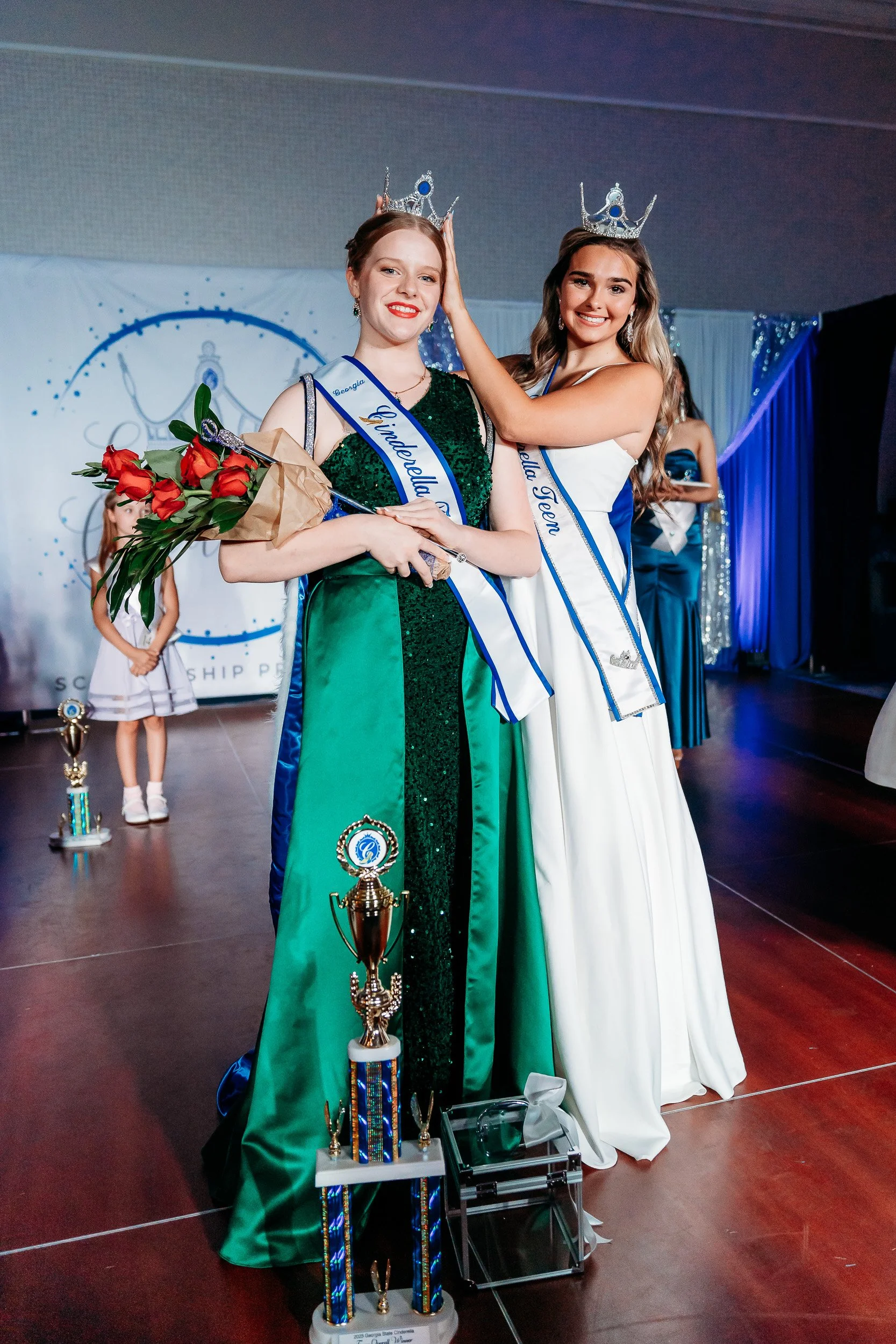 Two young women wearing crowns and sashes standing on stage during a pageant. One is holding a bouquet of flowers, and there are trophies in front of them. The woman on the left is wearing a green dress, and the woman on the right is wearing a white 