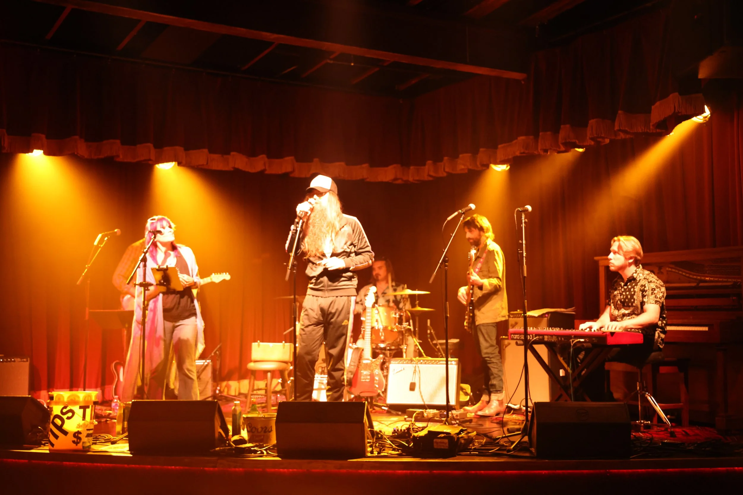 A musical performance on stage with five musicians, including singers, guitarists, a keyboardist, and a drummer, under warm stage lighting with a wooden backdrop and curtains.