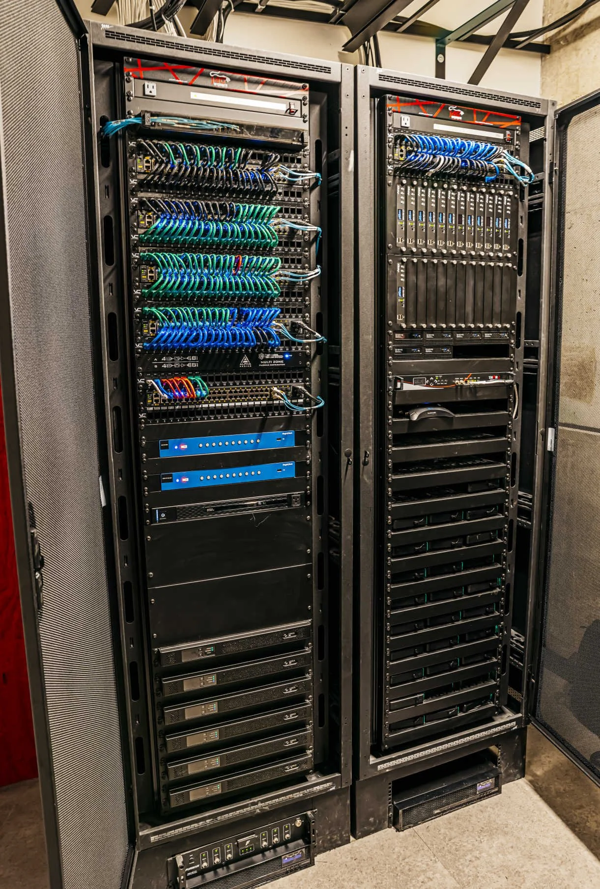 Two large black server racks with network equipment, cables, and electronic devices inside, situated in a server room.