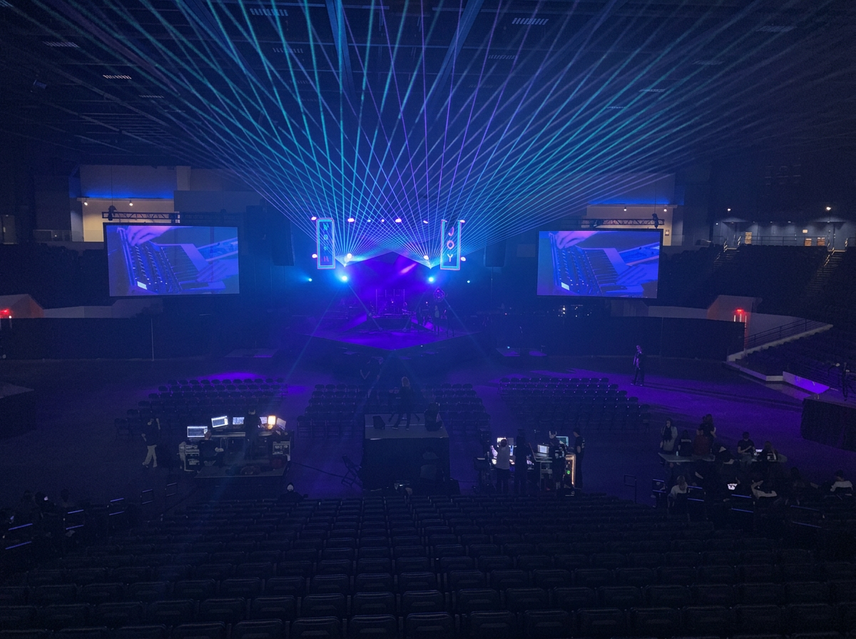 Empty indoor concert venue with stage, large screens, and laser light show in purple and blue colors.