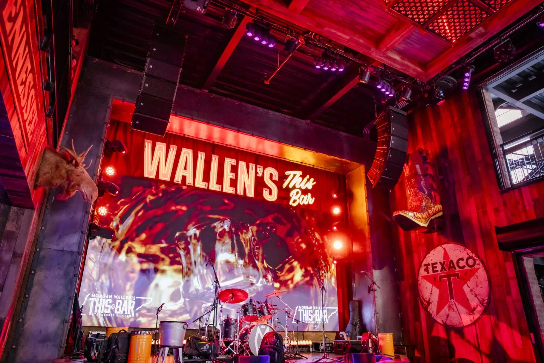 Stage setup at Wallen's This Bar with musical instruments, large screens, red and purple lighting, wood paneling, and mounted decorative animals.