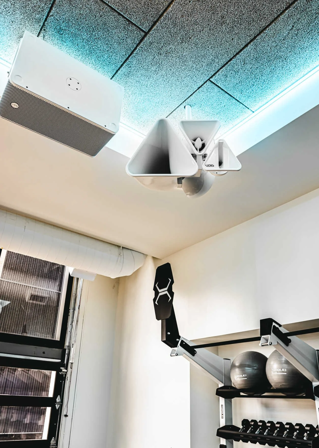 Gym inside with a white ceiling with acoustic tiles, a white speaker, a white hanger with two empty containers, a black wall-mounted resistance band anchor, and black exercise equipment including exercise balls and a dumbbell rack.