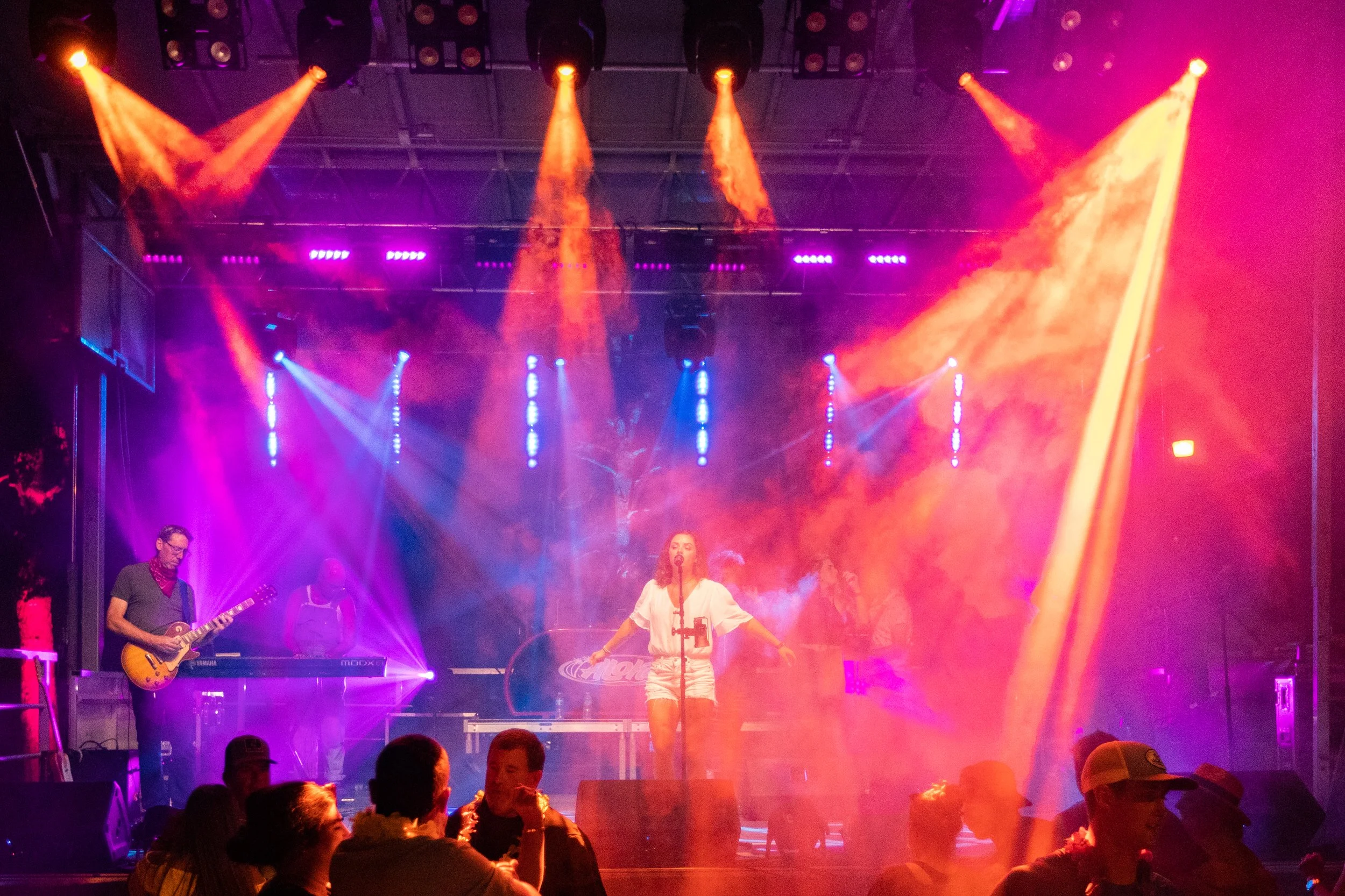 A live music performance on stage with colorful purple, orange, and yellow lighting effects, featuring a female singer at center, a guitarist on the left, and other band members in the background. The audience is visible in the foreground.