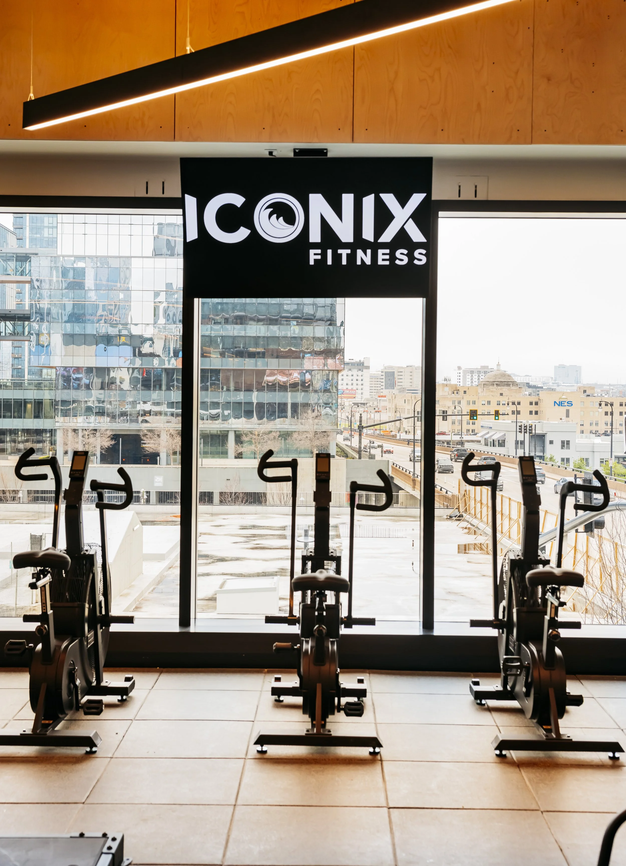 Indoor gym with exercise bikes positioned near large windows, cityscape visible outside, and a black sign with white text reading 'ICONIX FITNESS' hanging inside.