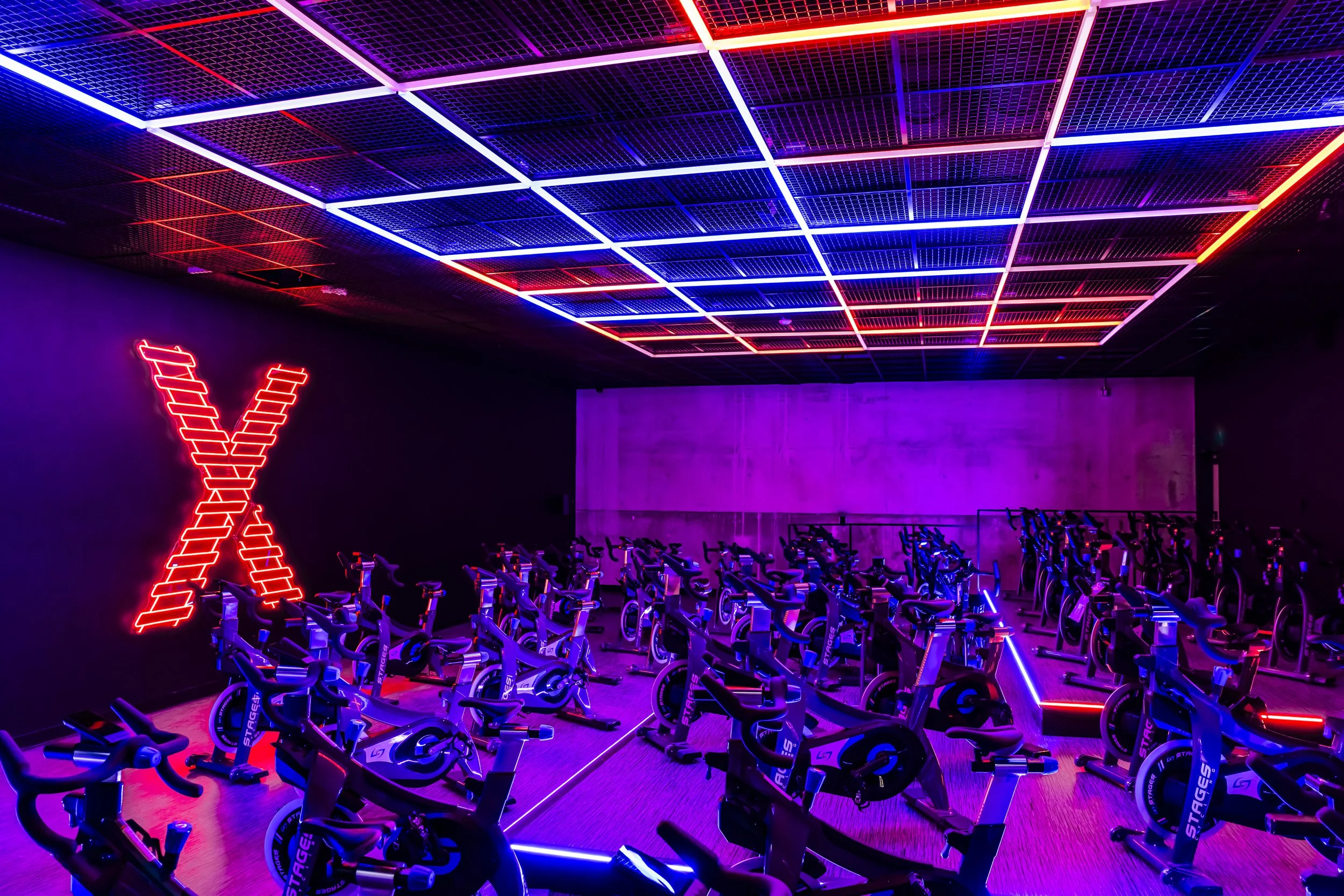 A dimly lit room with rows of stationary exercise bikes illuminated by vibrant neon purple, red, and blue lights on the ceiling. A large red neon letter X is on the wall to the left.