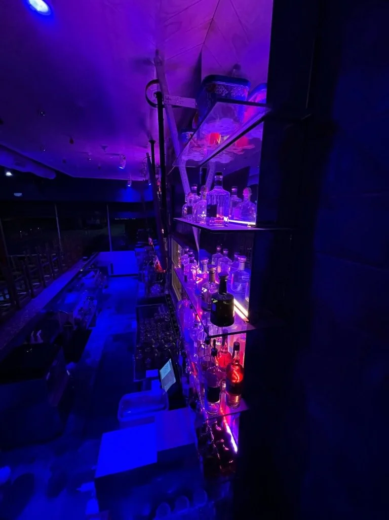 Bar counter with shelves of liquor bottles illuminated by purple and blue lighting in a nightclub or bar setting.