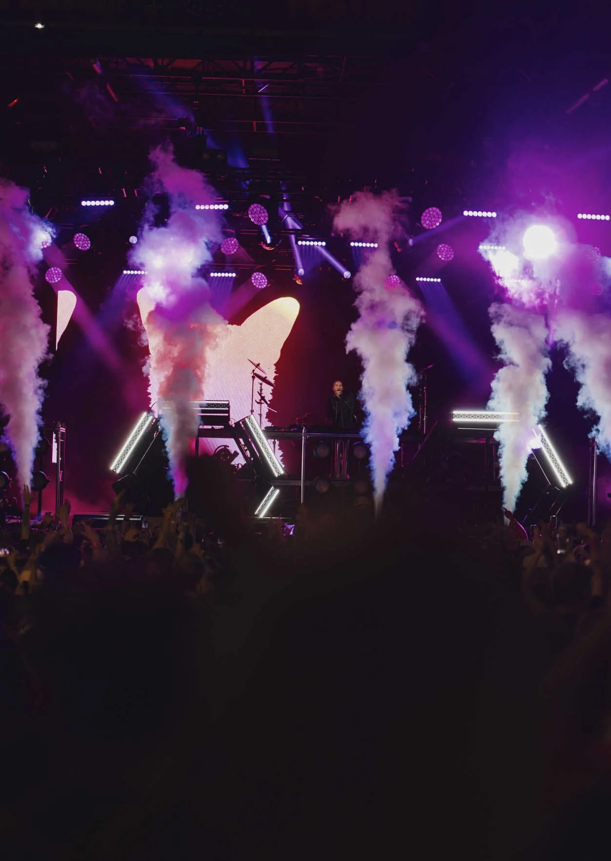 A live music concert stage with purple and white smoke effects, bright lights, and a DJ or performer at the center. Audience members' hands are visible at the bottom of the image.