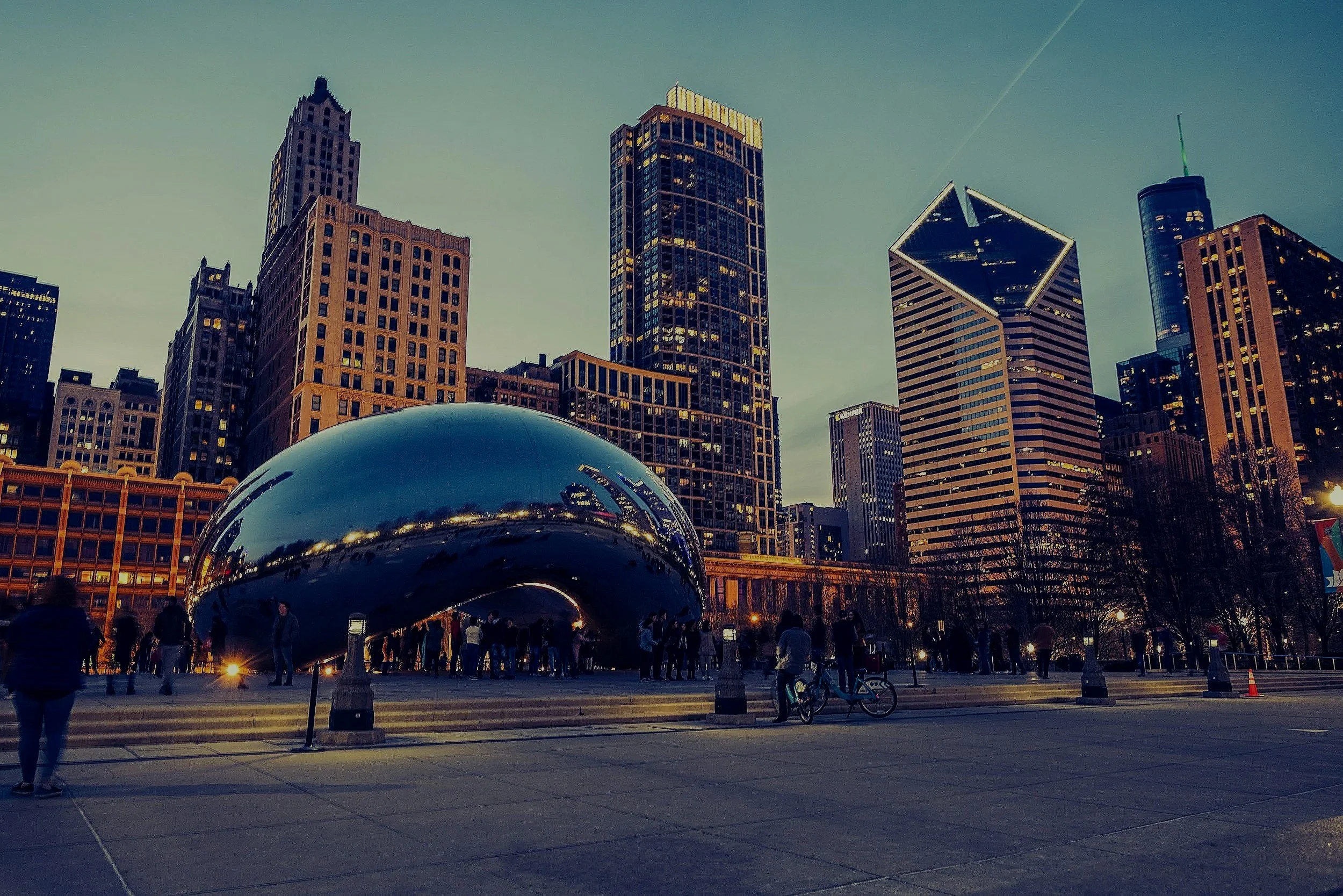 A view of Chicago's Millennium Park during dusk, featuring the Cloud Gate sculpture in the foreground and the city's skyline with tall buildings in the background.