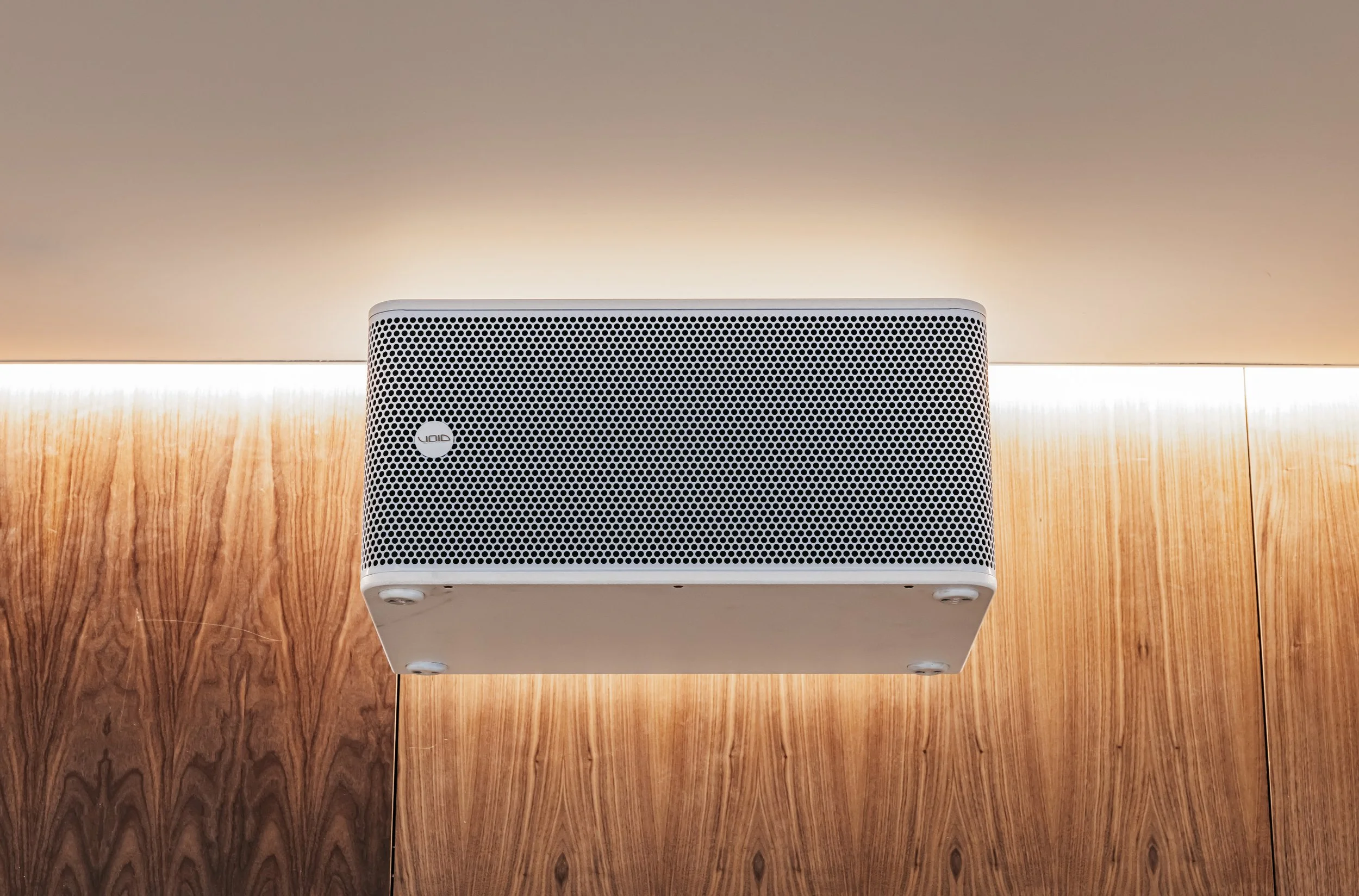 A mounted white speaker with a perforated front panel on a wooden wall with warm lighting.