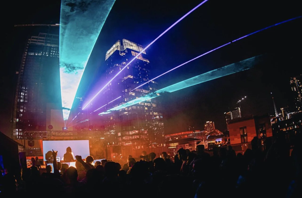Nighttime outdoor music event with a crowd, laser lights, a DJ booth, and city buildings in the background.