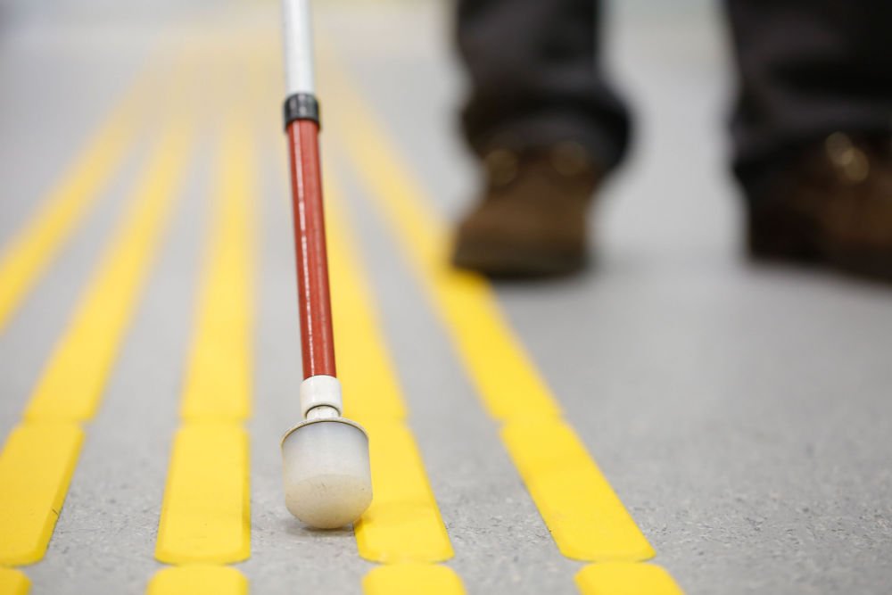 A white mobility cane against a yellow crosswalk. The user's brown shoes and pants are blurred in the background.