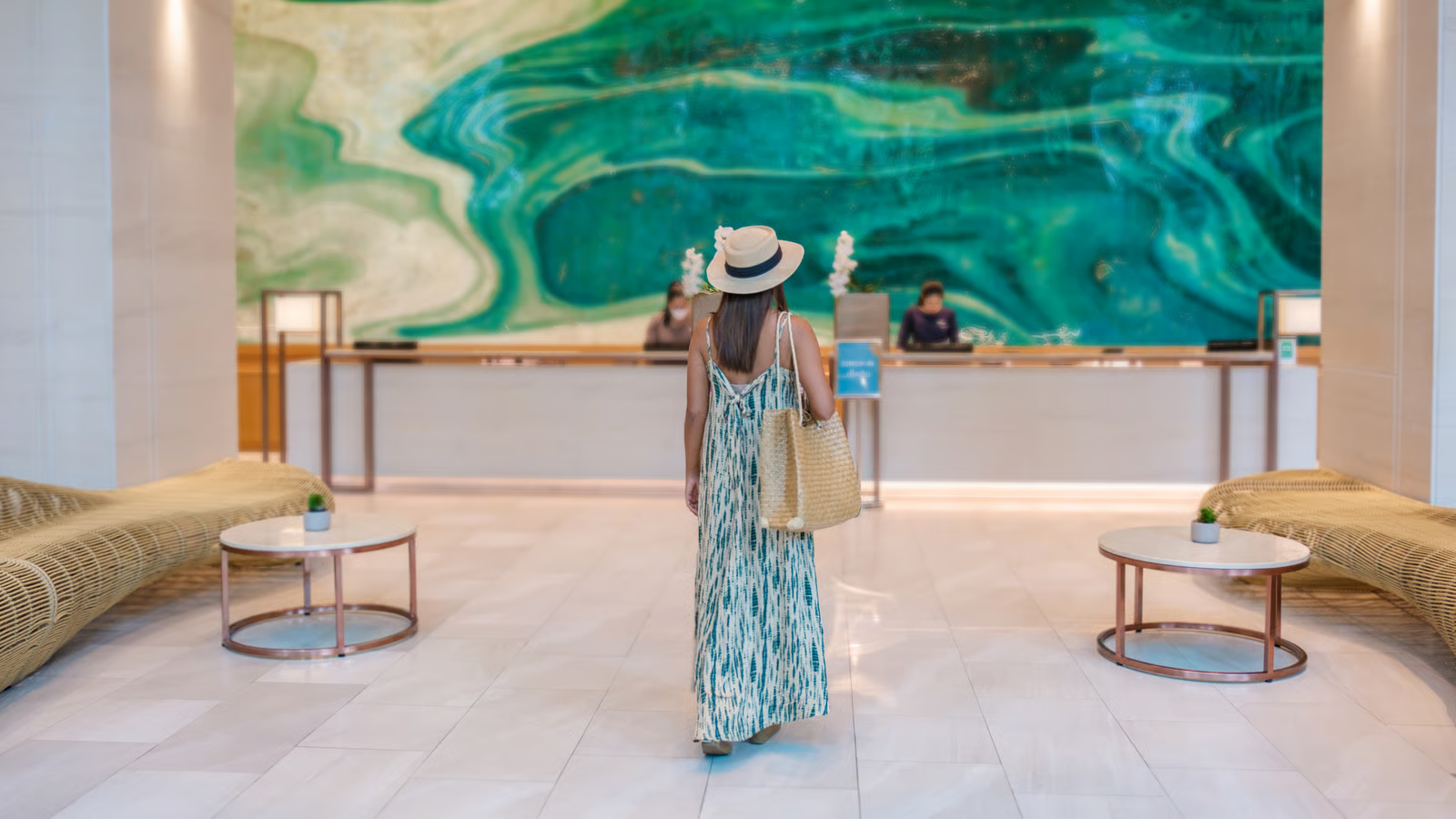 Woman in a straw hat and patterned maxi dress walks towards a hotel reception desk, with a large green abstract mural behind it and lounge seating on either side of the lobby.