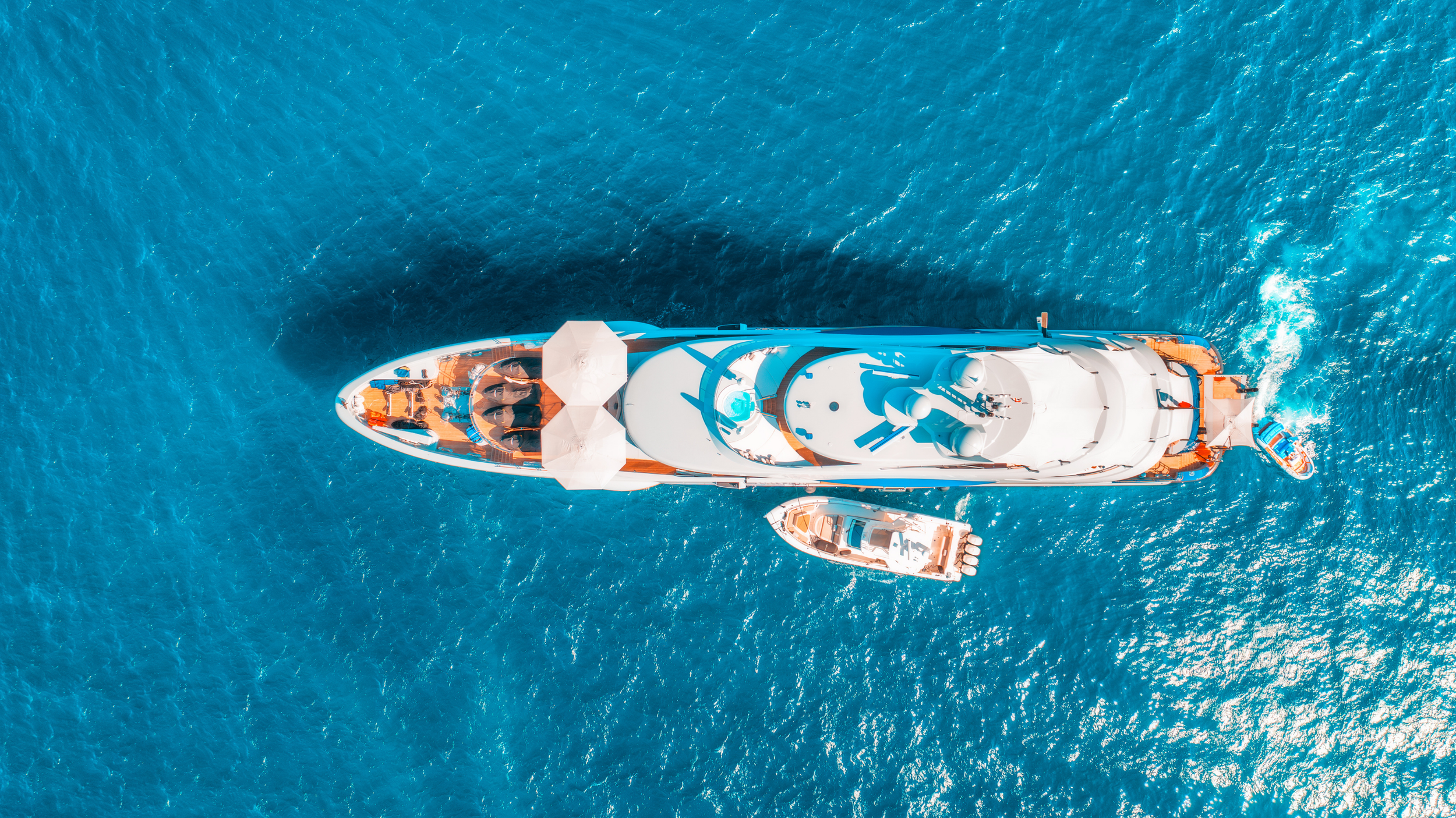 An aerial view of a large white luxury yacht with a smaller boat next to it, floating in the blue ocean.