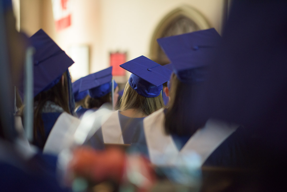 Students donning caps and gowns, sit waiting for their names to be called during the graduation ceremony. The photograph is captured at eye-level through rows of seated students, offering an interesting perspective and creating a sense of being immersed in the crowd.