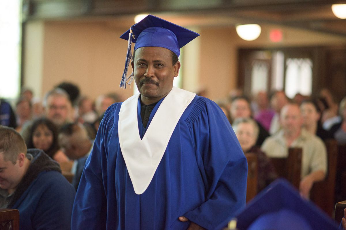 A student stands, preparing to approach the stage. The warm smile on the graduand’s face evokes a sense of pride and anticipationt. A shallow depth of field is used to bring the student into focus and softly blur the seated guests in the background.