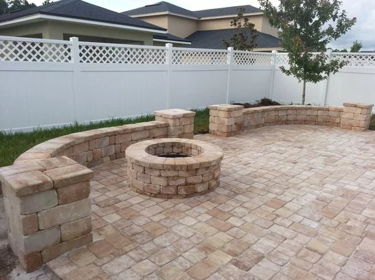 Backyard area with built-in curved seating around a brick fire pit, enclosed by a white fence with a tree in the corner.