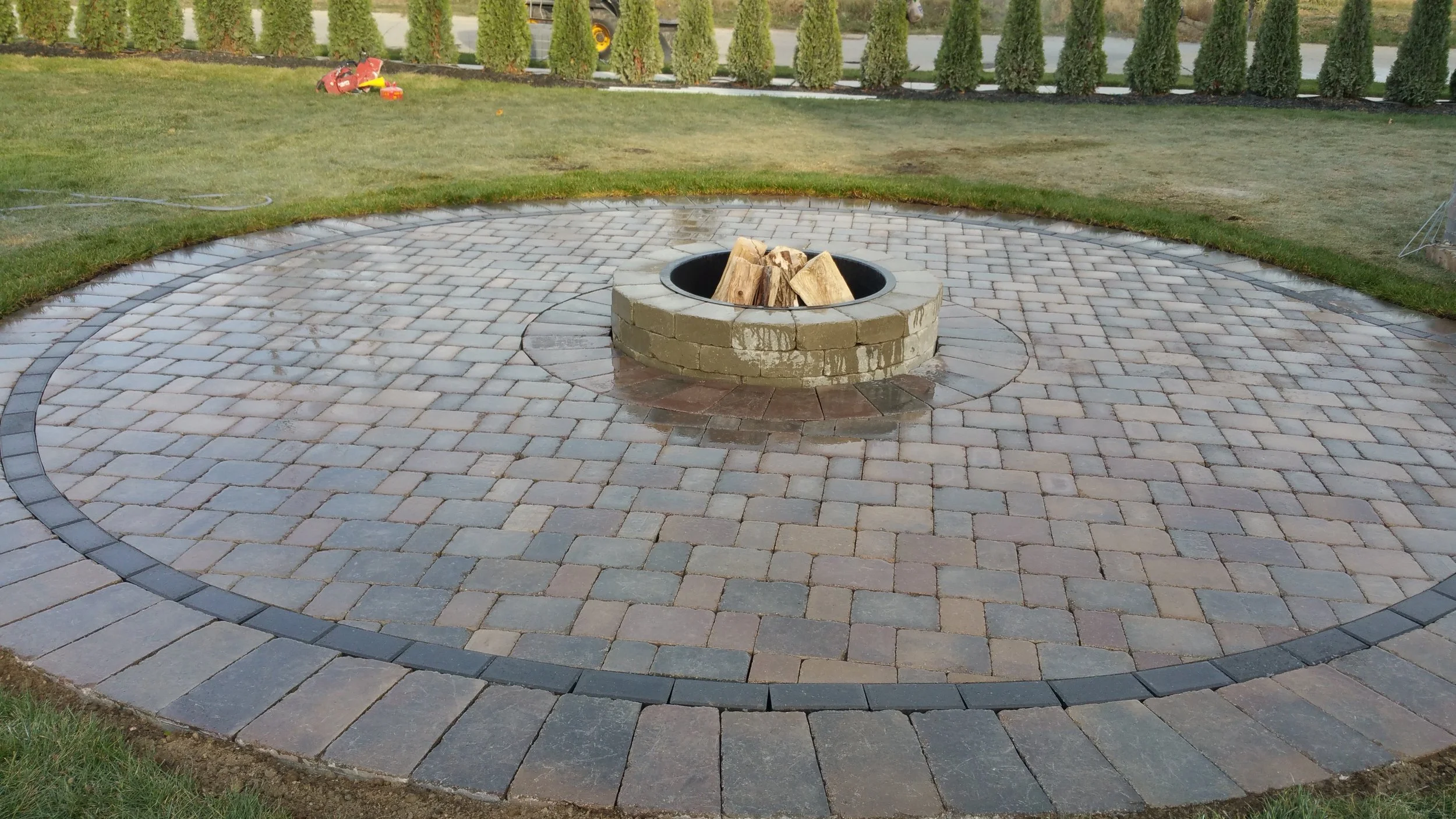 A newly paved circular brick area with a central fire pit filled with cut firewood, surrounded by a grassy yard and a row of tall, trimmed bushes.