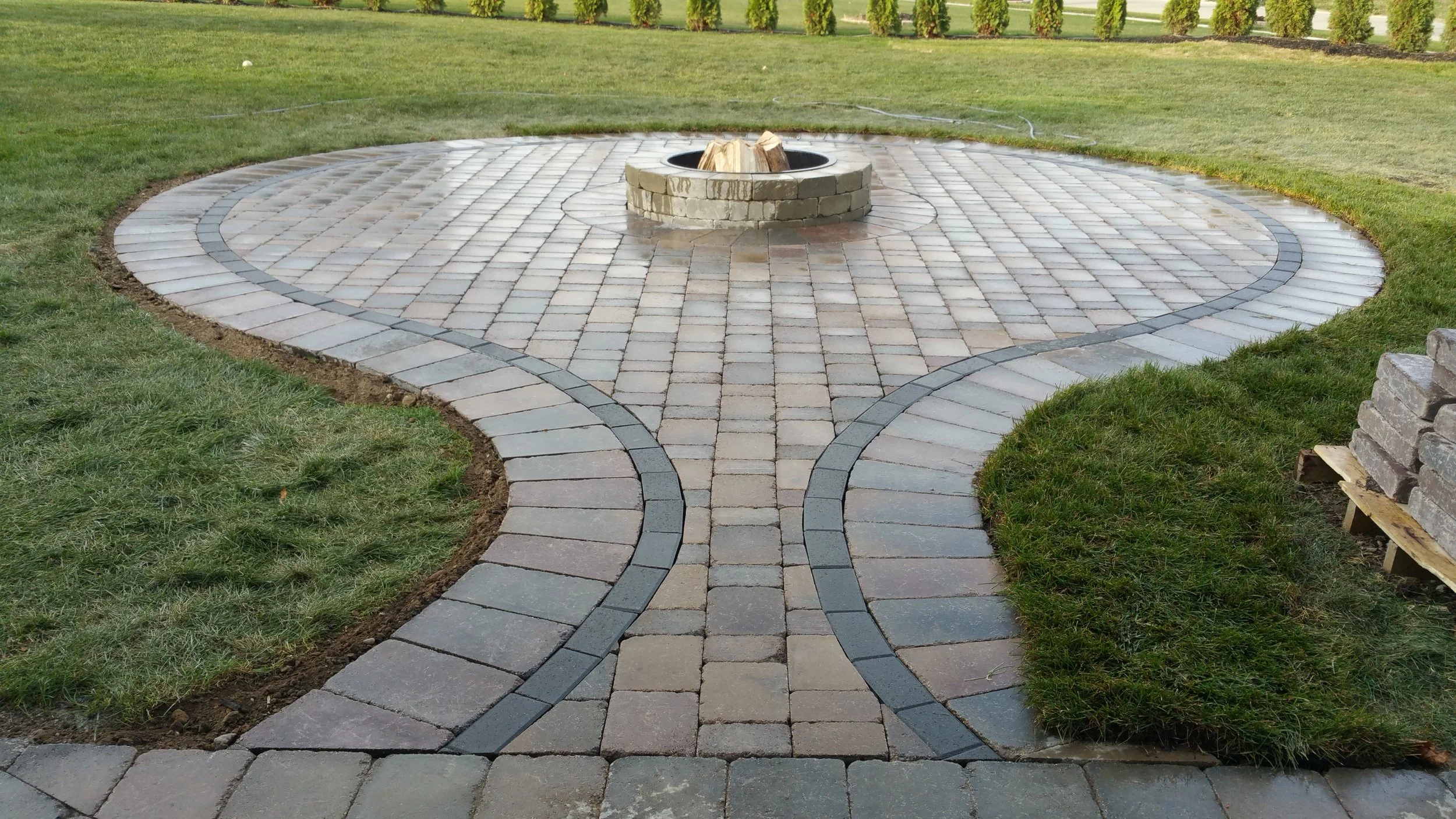 A landscaped outdoor area with a circular fire pit made of bricks, surrounded by paved pathways and green grass on either side.