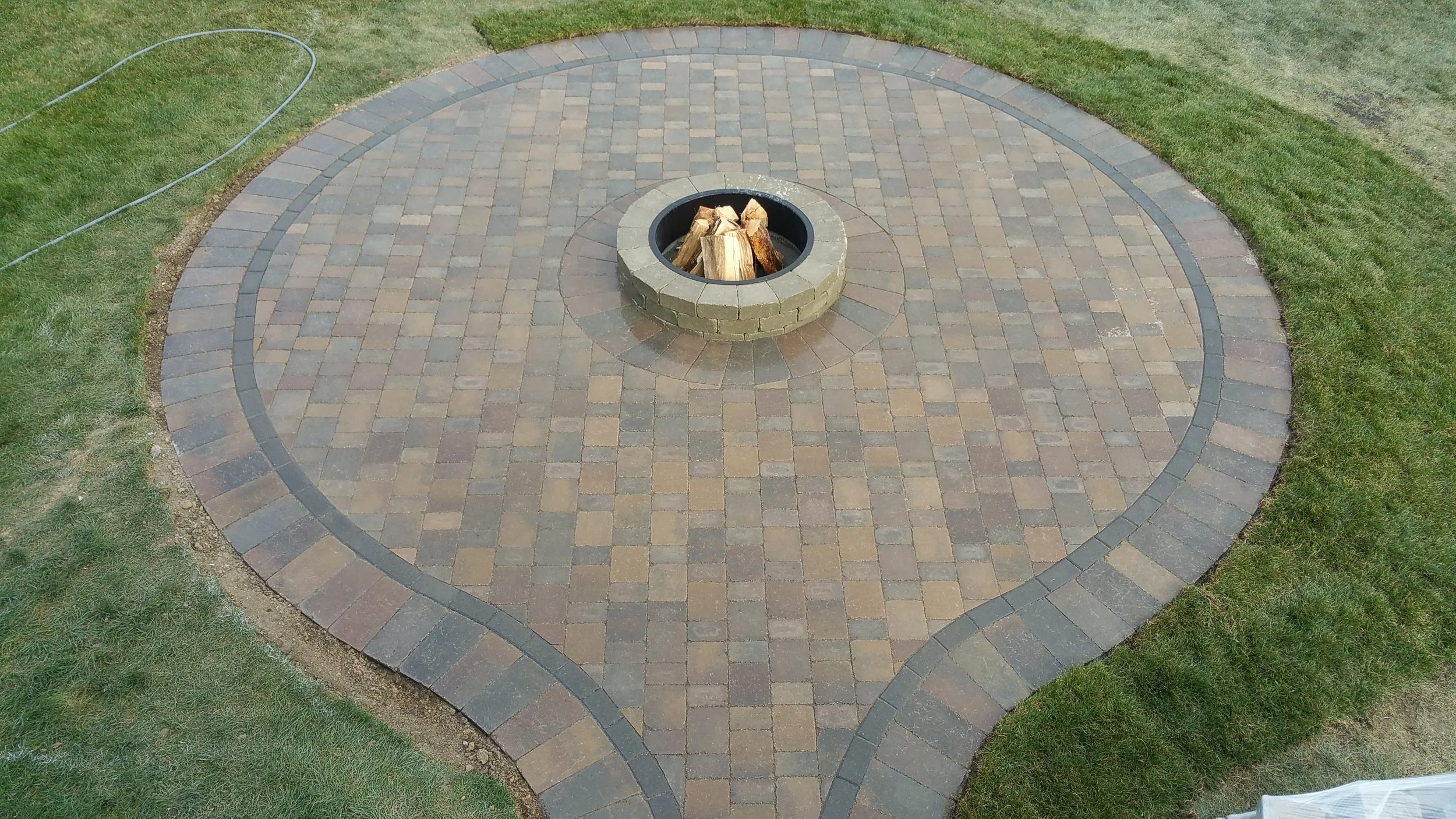 A  brick landing with a fire pit in the center containing logs, surrounded by grass.