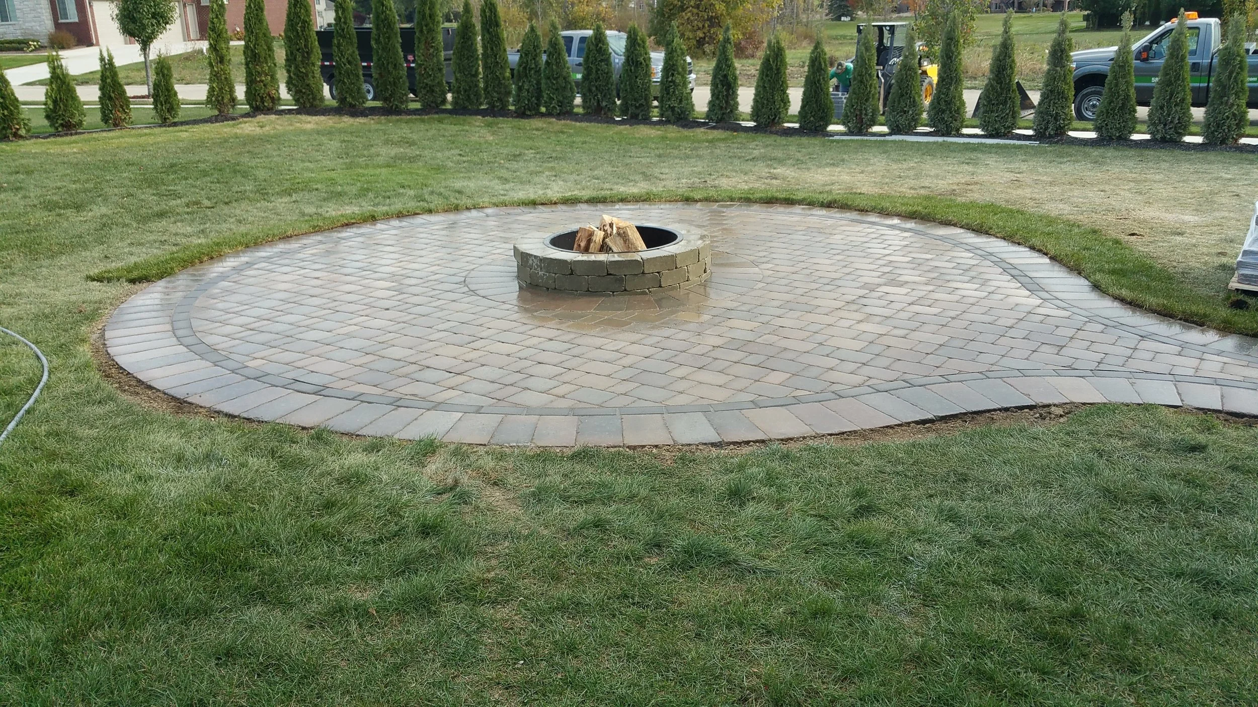 A newly paved circular stone area with a fire pit at the center, surrounded by a green lawn and a row of tall, slender evergreen trees in the background.
