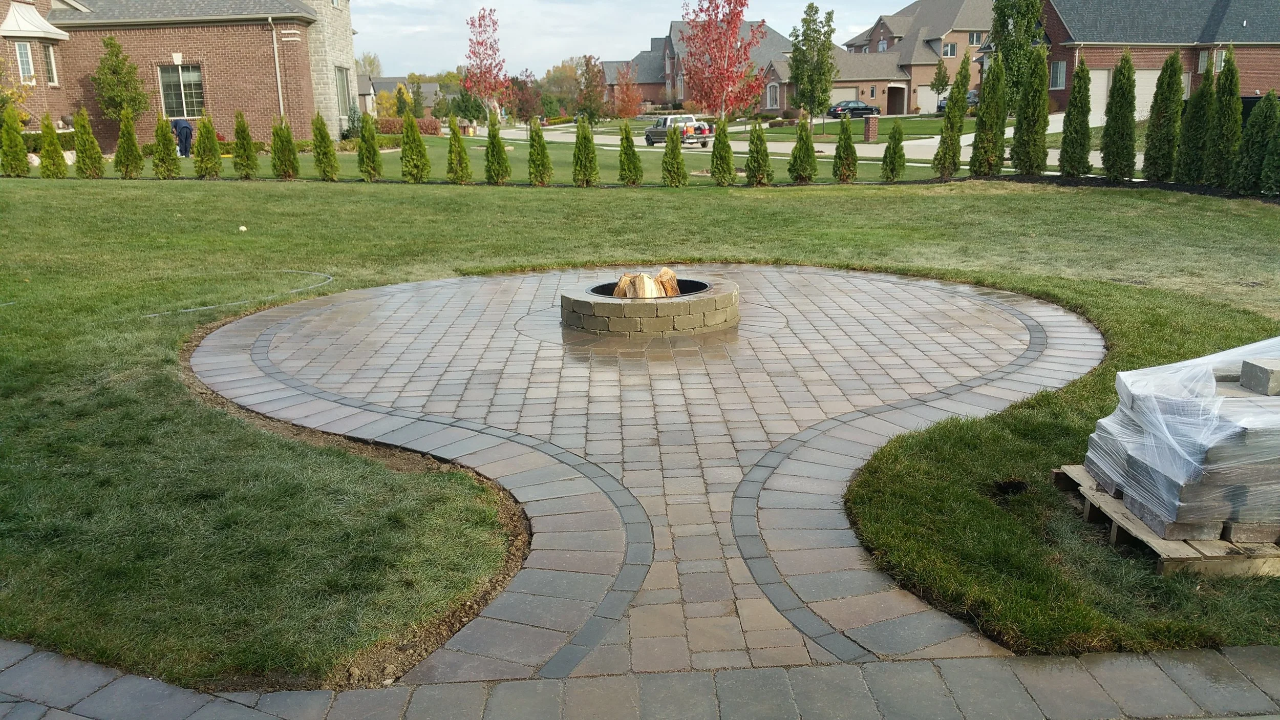 Backyard with a round paved area and a small fire pit at the center, surrounded by well-maintained grass and a row of tall, narrow evergreen trees in the background, with houses and a street further behind.