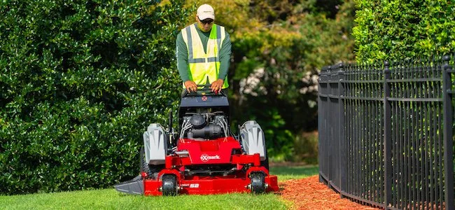 A man operating a red lawn mower on a grassy lawn next to a black metal fence and green bushes.
