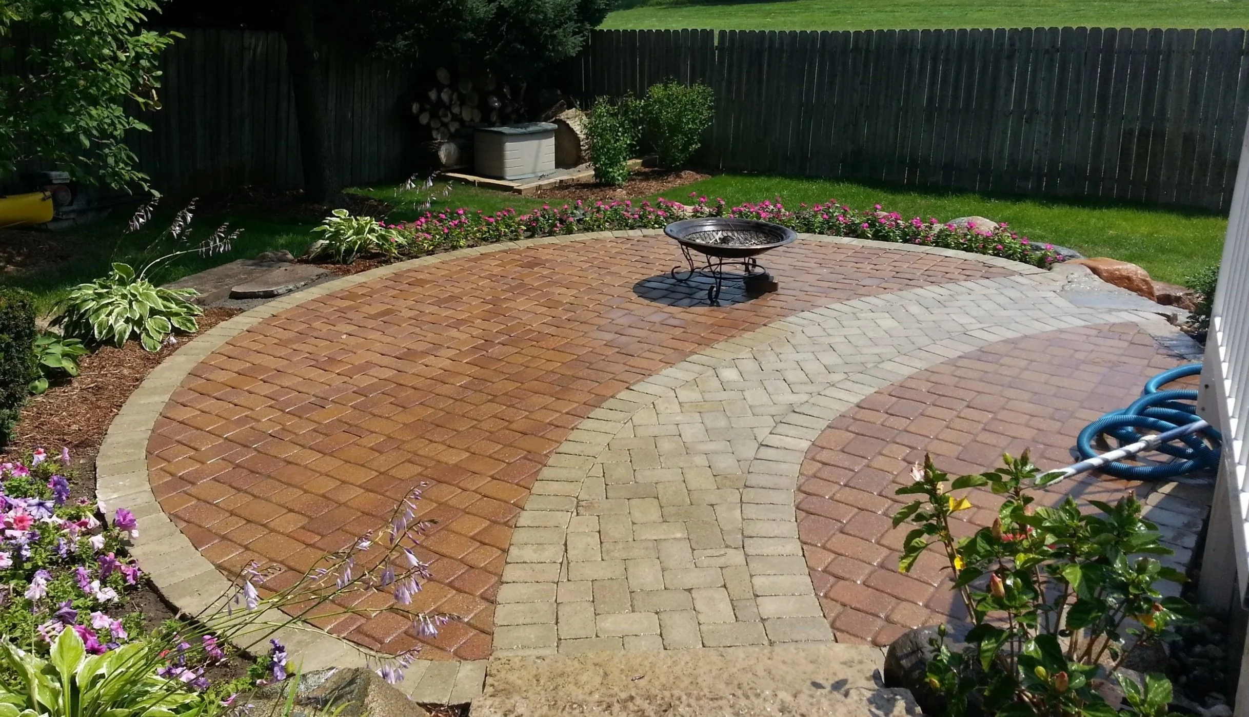 Backyard patio with brick pavers, fire pit in the center, surrounded by flowers and green grass, fenced perimeter, in sunlight.