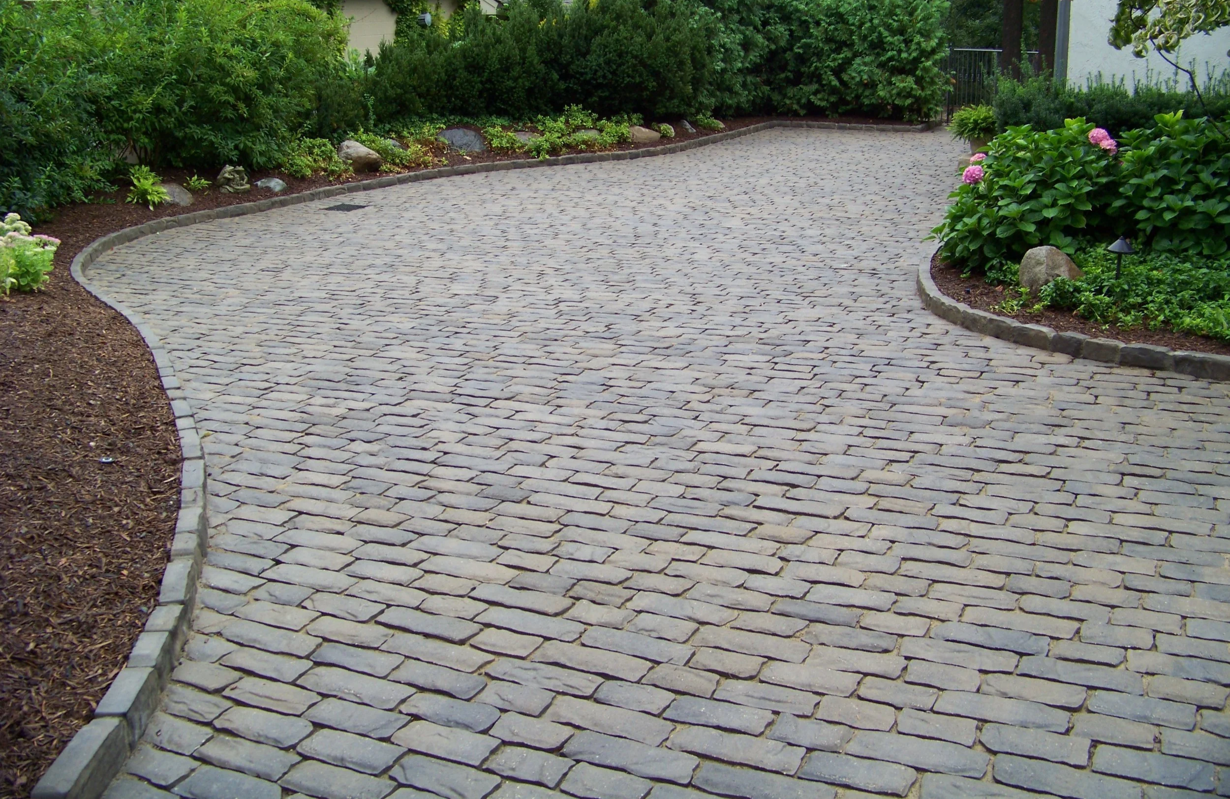 A curved stone paver driveway bordered by garden beds with green bushes, flowers, rocks, mulch, and landscape lighting.