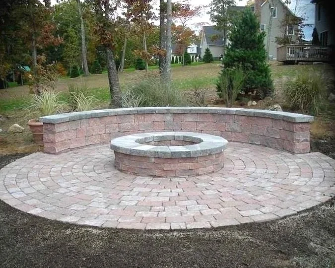 A circular brick patio with a central fire pit, surrounded by a low curved brick wall, situated outdoors in a yard with trees and houses in the background.