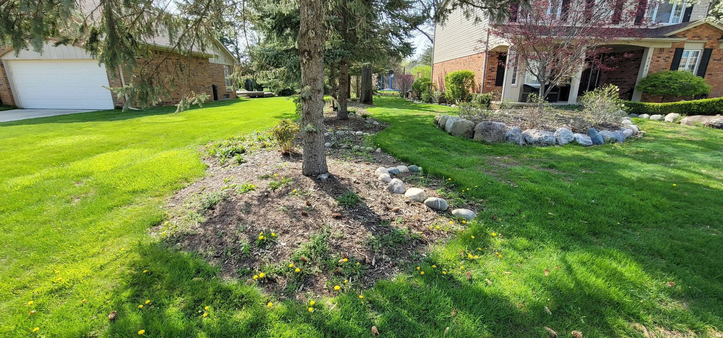 A front yard with green grass, trees, flower beds with rocks, and a brick house with a porch.