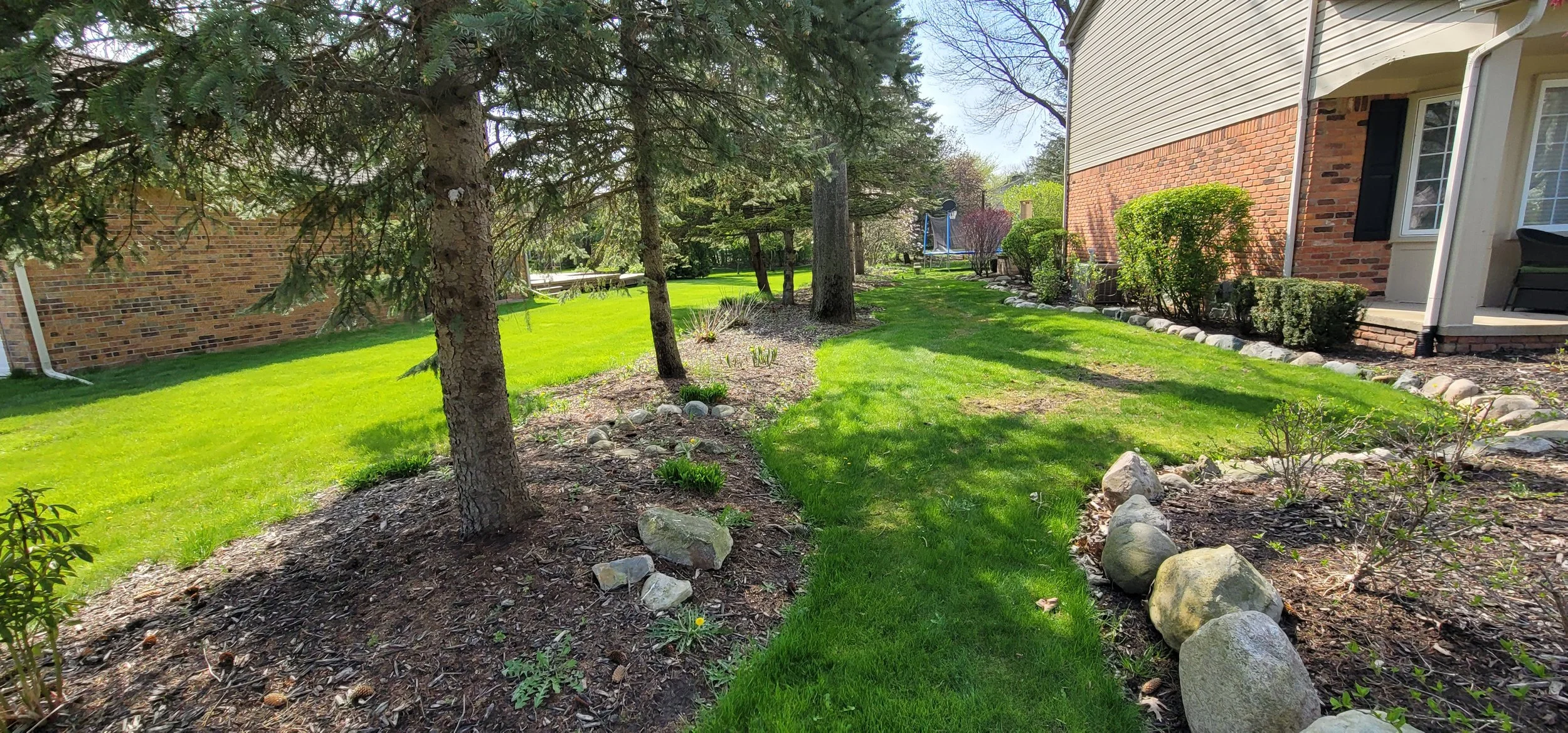 Backyard lawn with trees, shrubs, and flower beds near a brick house with a porch and black shutters.
