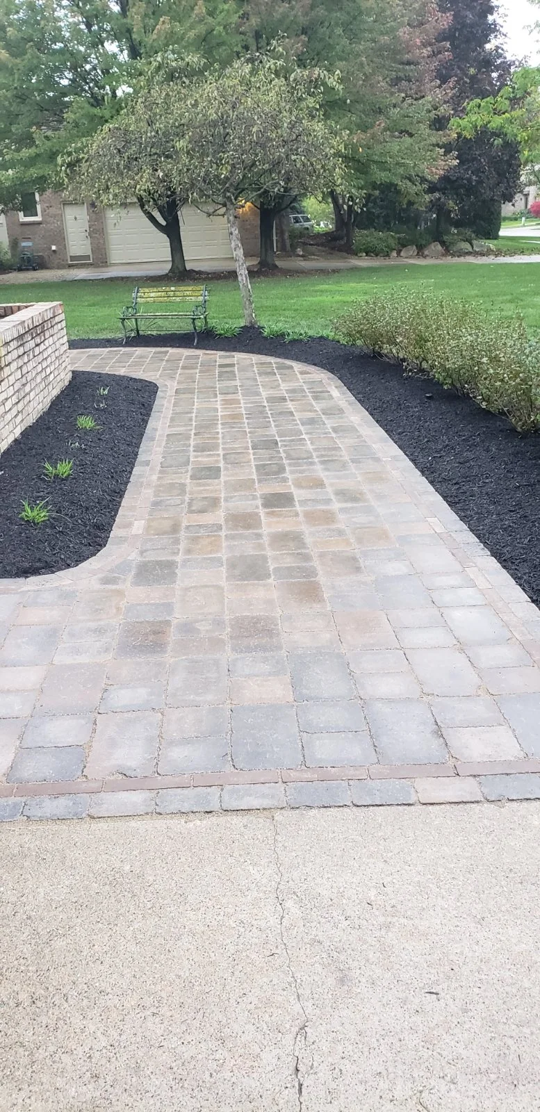 A paved walkway in a backyard with a brick wall on the left, a small tree, and a bench. There are bushes and trees in the background along with a grassy lawn.