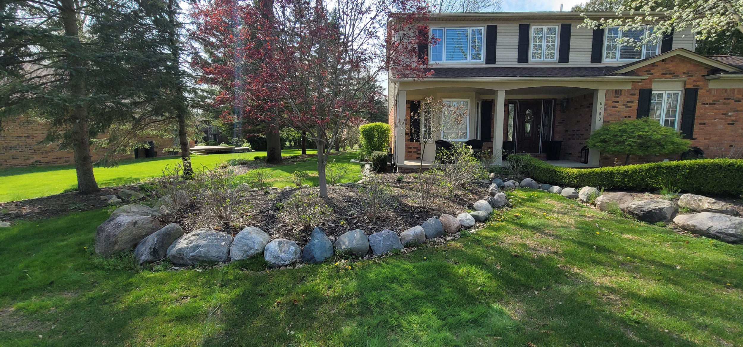 Front yard of a house with a garden bed surrounded by rocks, trees, green lawn, and a patio with chairs. The house has brick and light-colored siding with black window shutters.