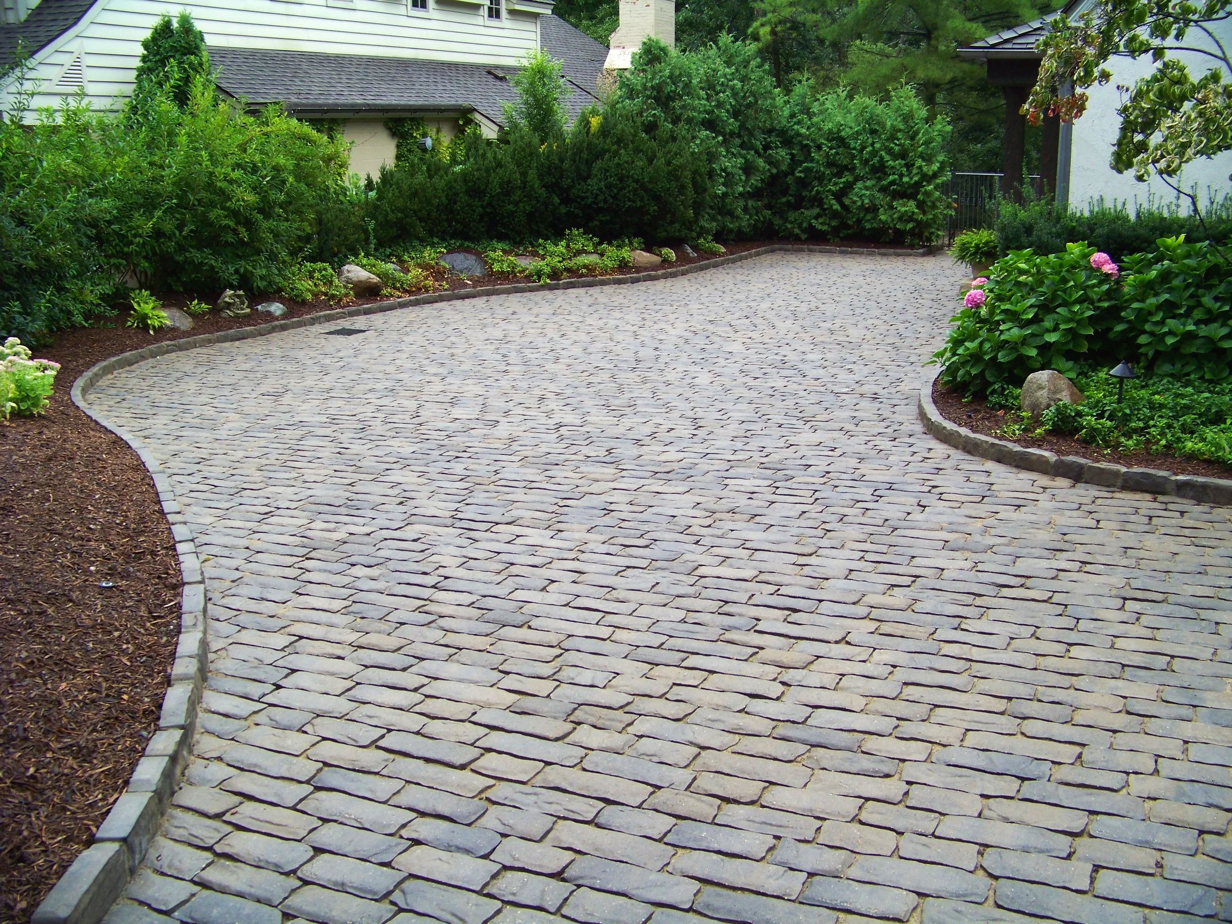 A paved driveway with interlocking stone tiles, bordered by flower beds with green bushes and colorful flowers, in a suburban yard.