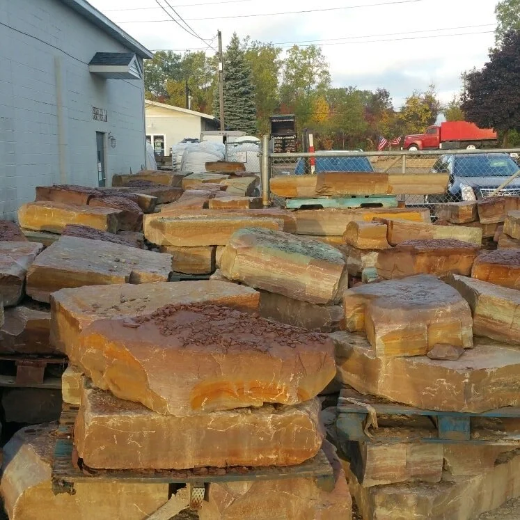 Stacks of large sandstone slabs outdoors near a chain-link fence, with trees, vehicles, and a building in the background.