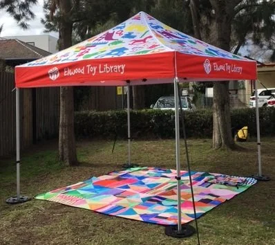A marquee set up in a park, 3 square metres, with colourful toy-themed top. A colourful picnic rug is on the grass beneath the marquee.