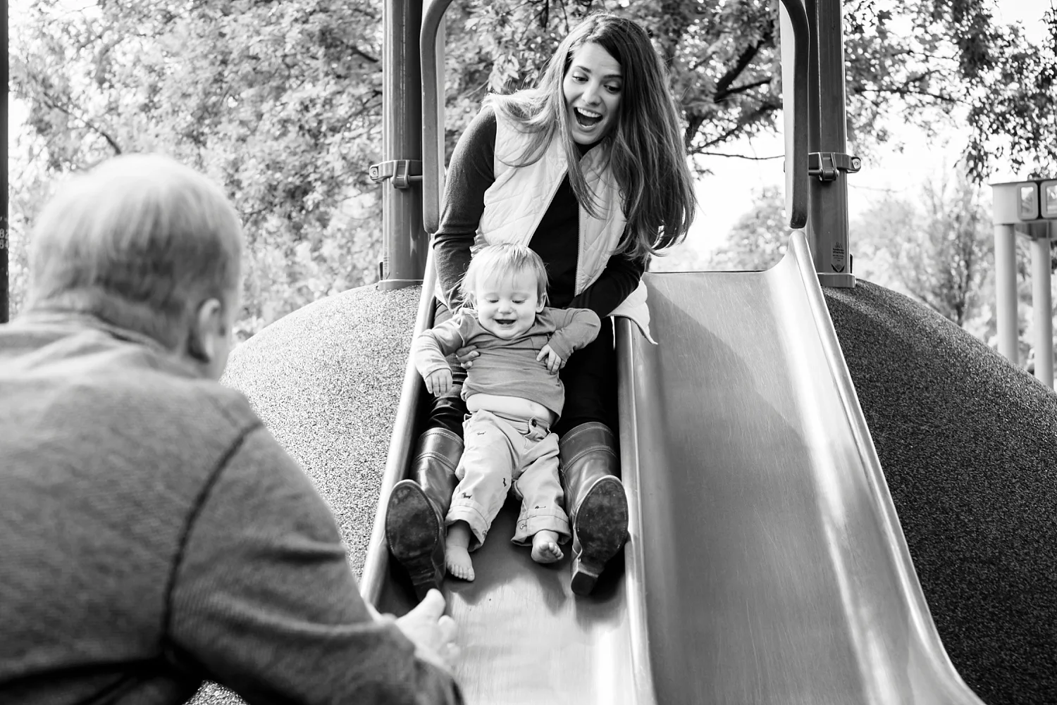A mom rides the slide with her toddler son in Denver, Colorado. Black and white family lifestyle photography with Kristiina Craven Photography.