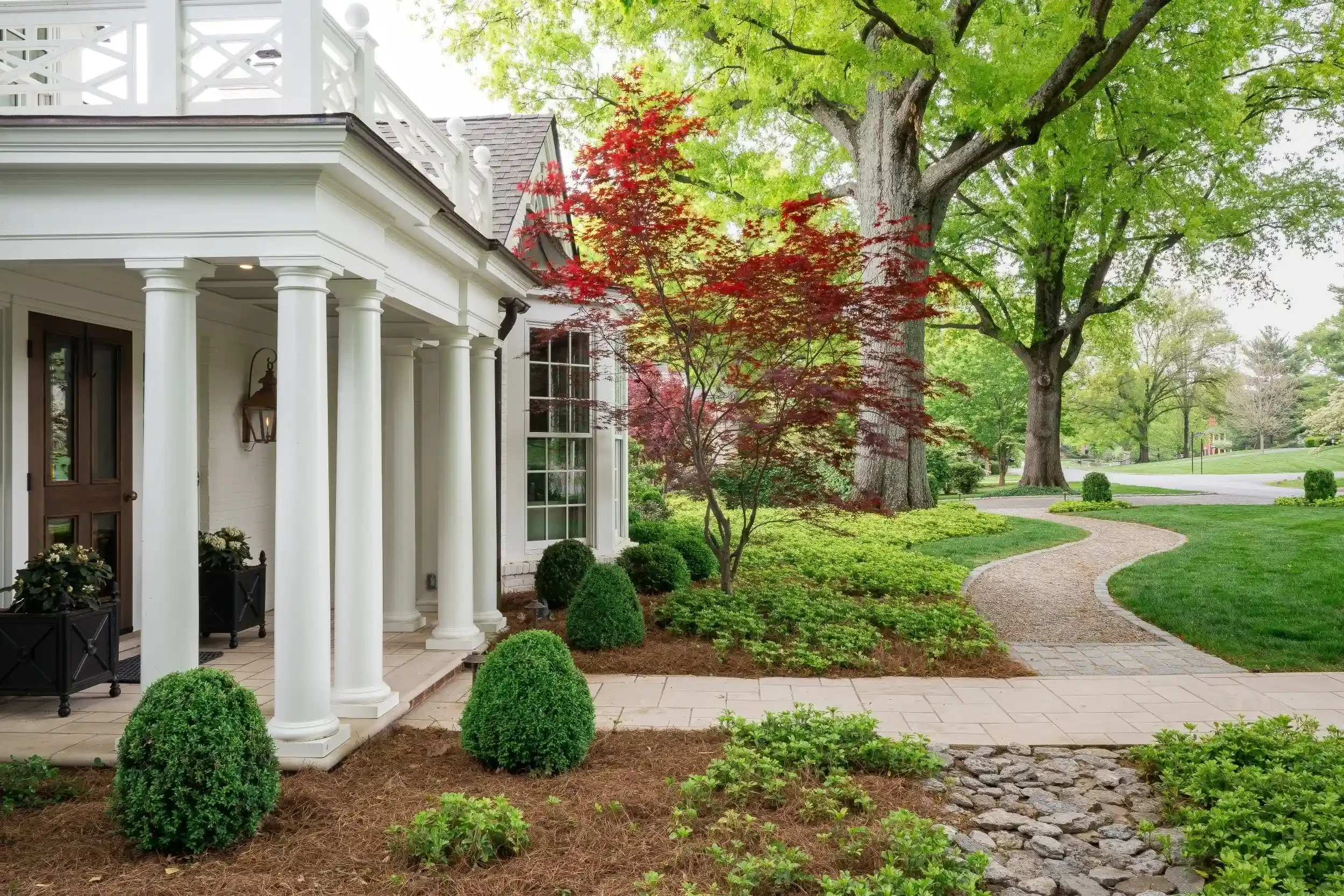 A front yard with a curved stone and gravel pathway, green grass, and neatly trimmed bushes. There are large trees with lush green leaves and a small red-leafed tree near a white house with classical columns and a porch. The scene is bright and well-maintained.