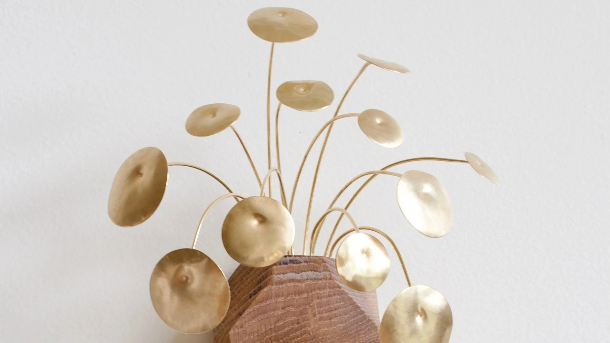 Cropped photo of handcrafted brass and wood wall sculpture, the brass leaves modeled after the Chinese Money Plant and look like gold coins against a white backdrop