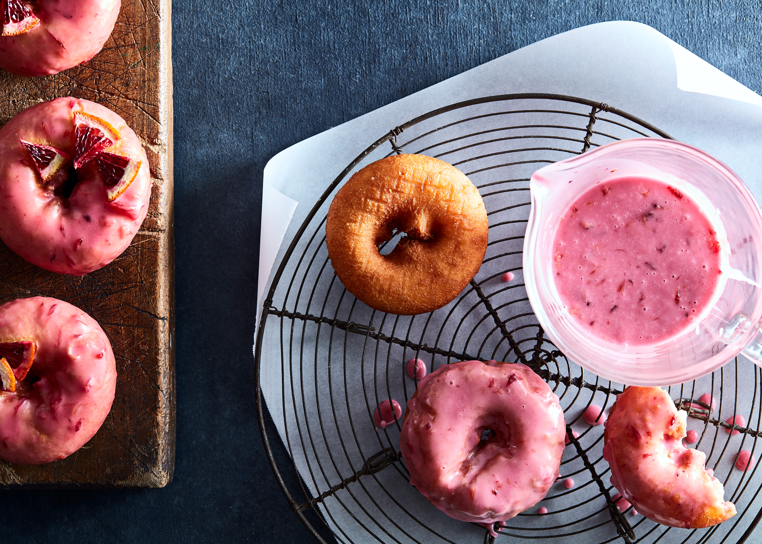 Pink blood orange glazed cake donuts on cooling rack with cup of icing for glazing.