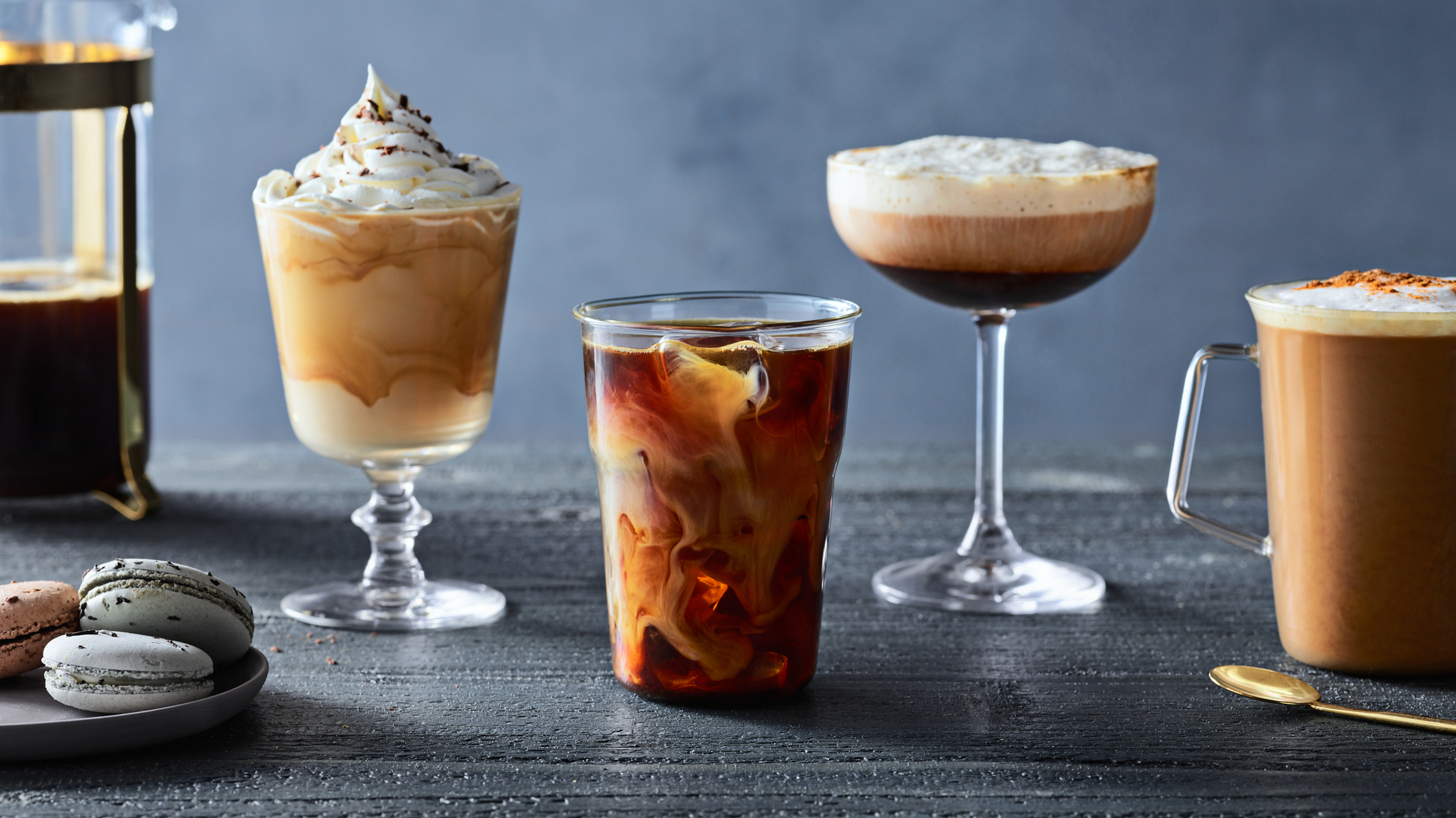 A lineup of various coffee drinks; iced coffee, espresso martini, hot coco, frozen coffee and plate of macarons.