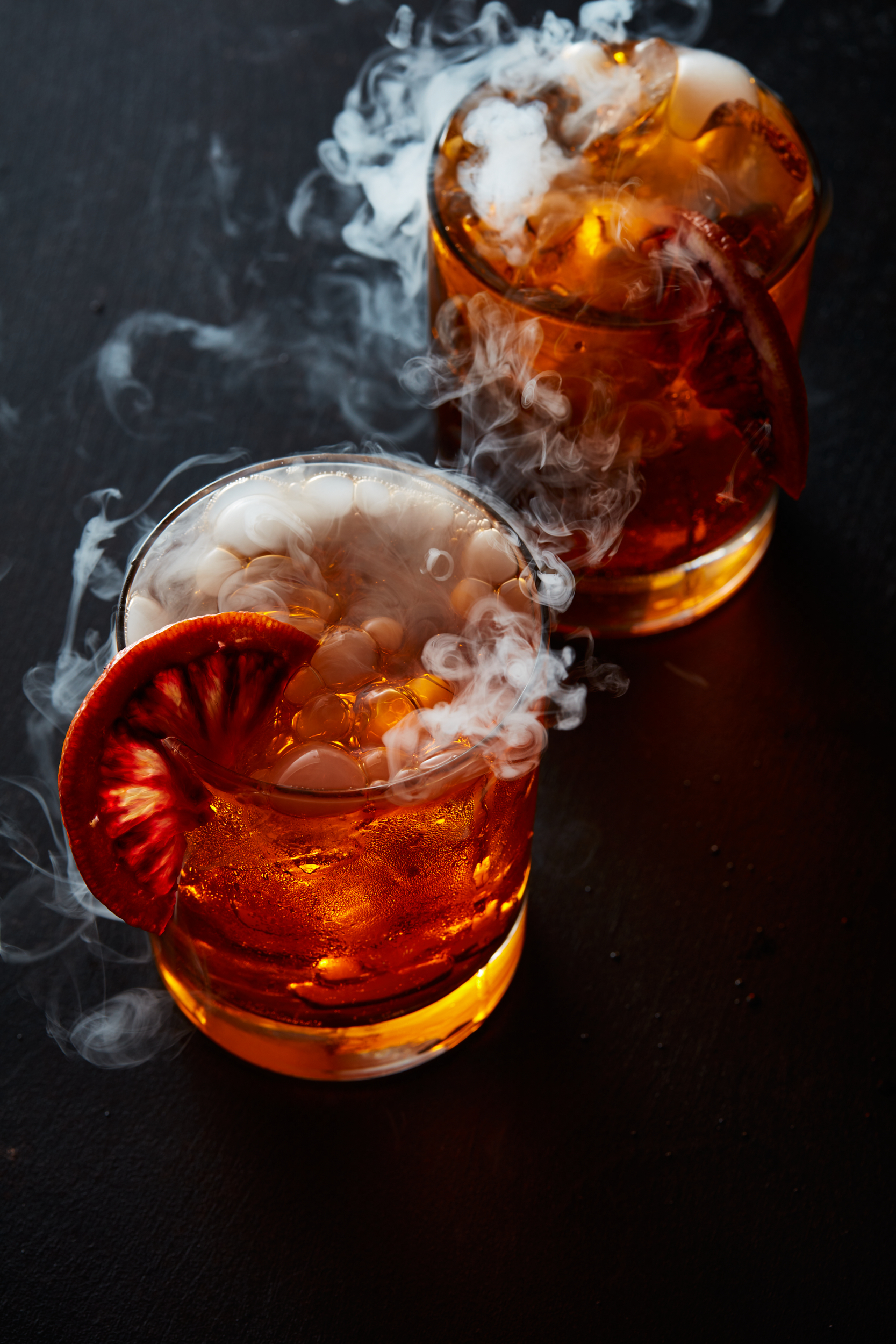 Two blood orange old fashioned cocktails with dry ice and smoke pouring out of the rims. Halloween themed cocktail photography.