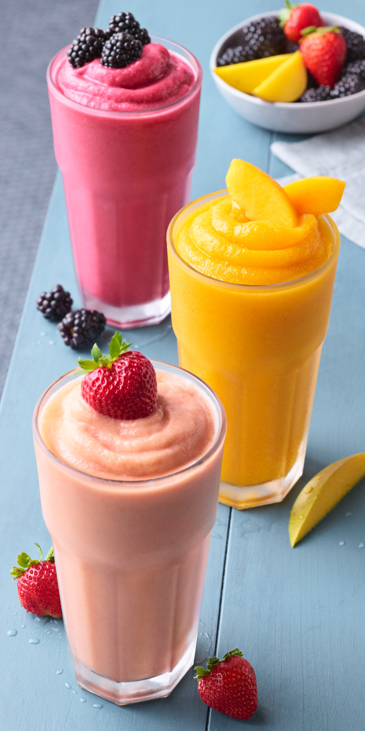 Three freshly made smoothies on a blue tabletop; strawberry, mango and blackberry.