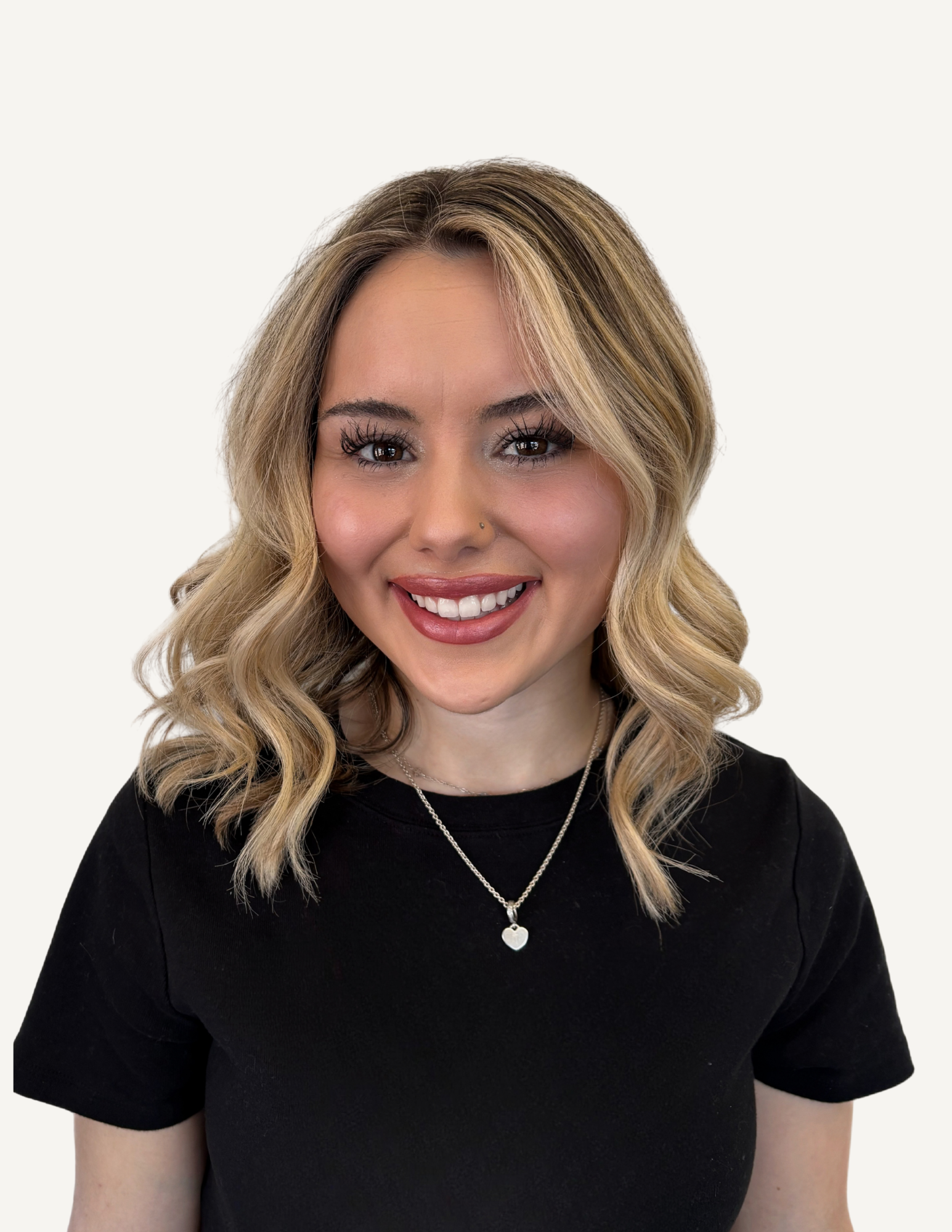 A young woman with long wavy blonde hair, wearing a black sleeveless top and a silver necklace, smiling at the camera indoors.
