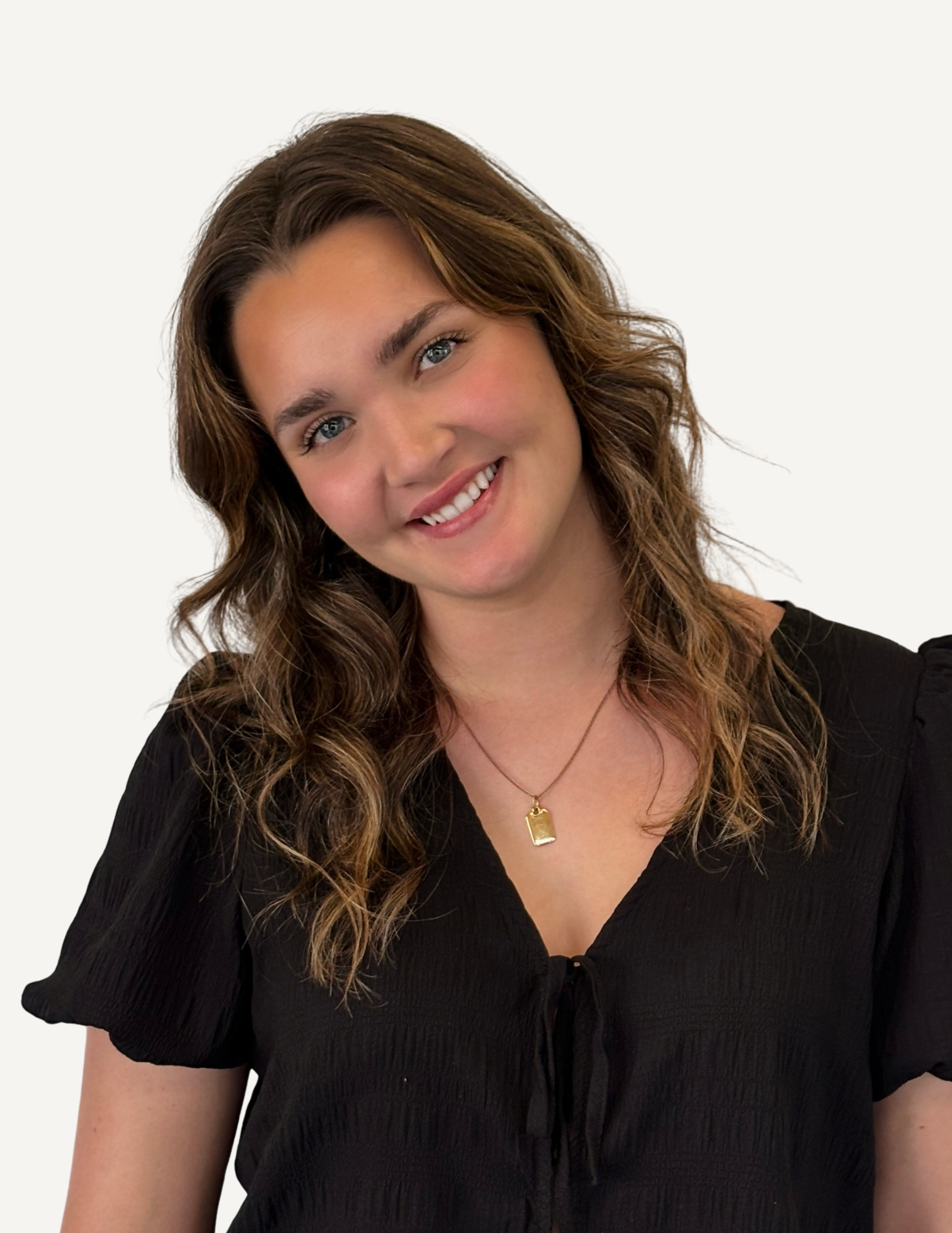 A young woman with wavy brown hair and blue eyes, smiling in a store setting, wearing a black top and layered necklaces, with shelves of products and a mirror in the background.