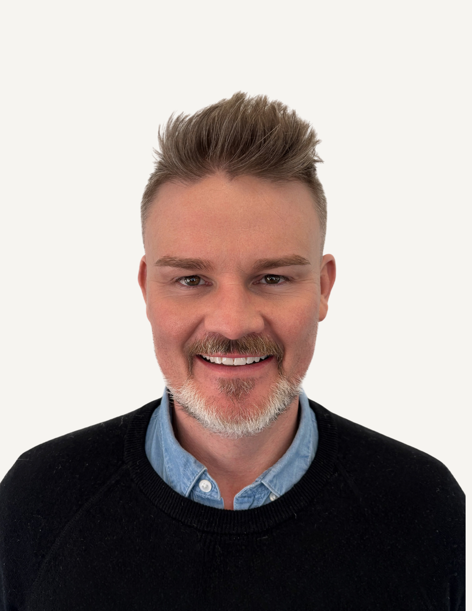 A smiling man with a beard and short, styled hair, wearing a black zip-up shirt, in an indoor setting with black furniture and mirrors in the background.