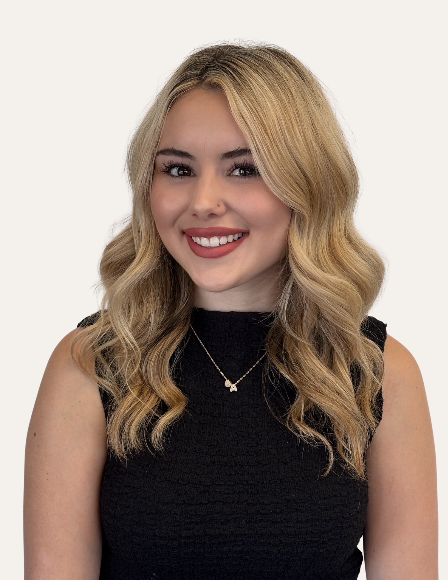 A young woman with long wavy blonde hair, wearing a black sleeveless top and a silver necklace, smiling at the camera indoors.